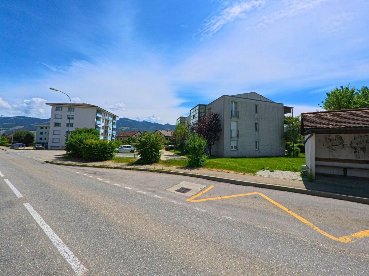In einem Wohnviertel von Riaz (1632, Kanton Freiburg) stehen moderne Mehrfamilienhäuser entlang einer ruhigen Strasse mit Bushaltestelle im Vordergrund und Blick auf das Bergpanorama.
