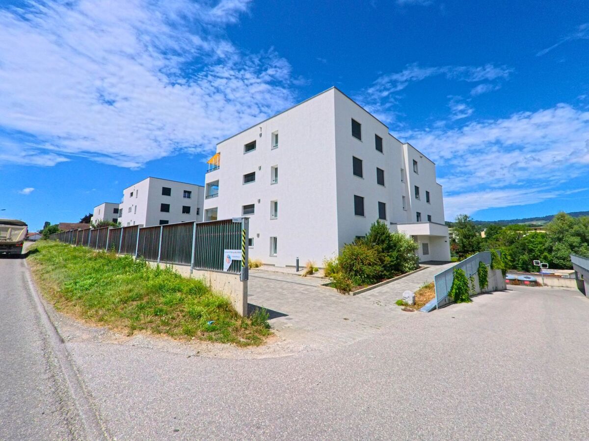 In Riaz (1632, Kanton Freiburg) reihen sich moderne, weisse Wohnbauten entlang einer ruhigen Strasse mit abfallender Zufahrt zur Tiefgarage und Blick auf die grüne Umgebung.