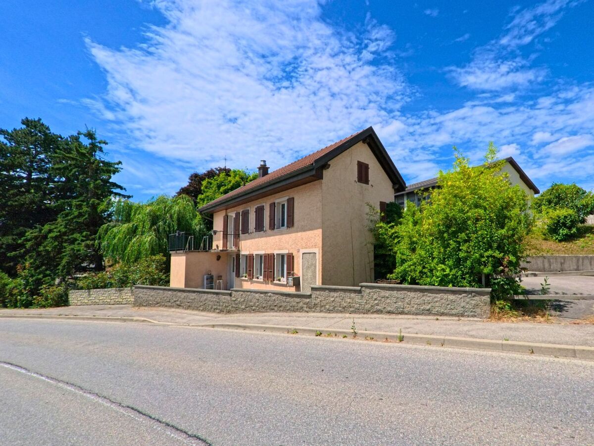 Ein hell verputztes Wohnhaus mit braunen Fensterläden steht in Riaz (1632, Kanton Freiburg) an einer leicht gebogenen Strasse, umgeben von Bäumen und Hecken.