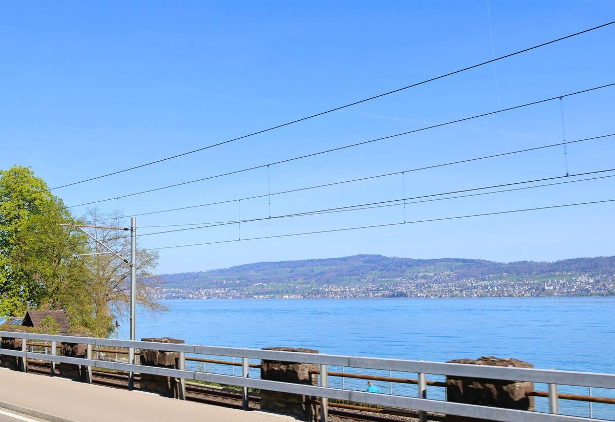In Richterswil, 8805 im Kanton Zürich, verläuft eine Bahnlinie direkt am Zürichseeufer entlang und eröffnet den Blick auf das gegenüberliegende Seeufer mit Hügeln.
