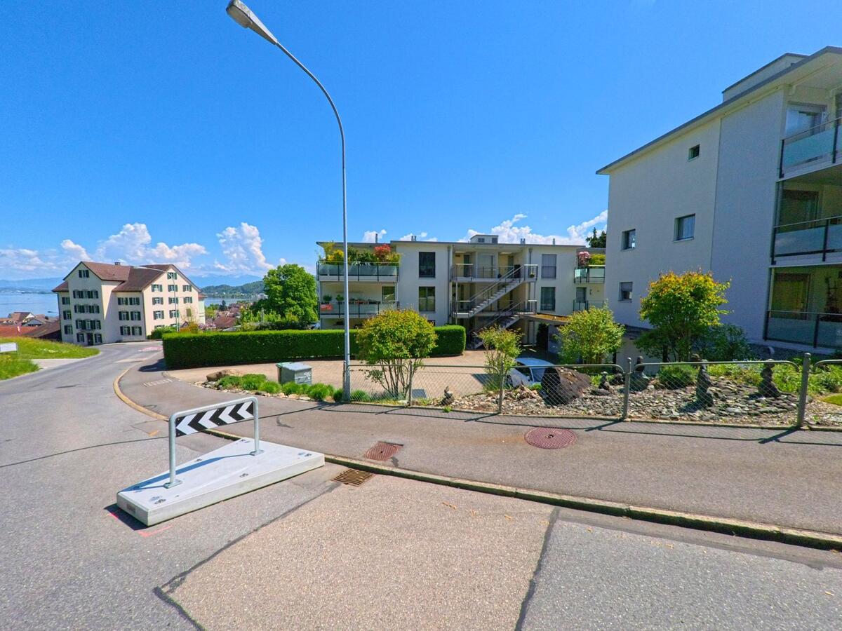  In Richterswil, 8805 im Kanton Zürich, verläuft eine Wohnstrasse mit modernen Mehrfamilienhäusern und Seesicht, flankiert von einer kleinen Verkehrsinsel.