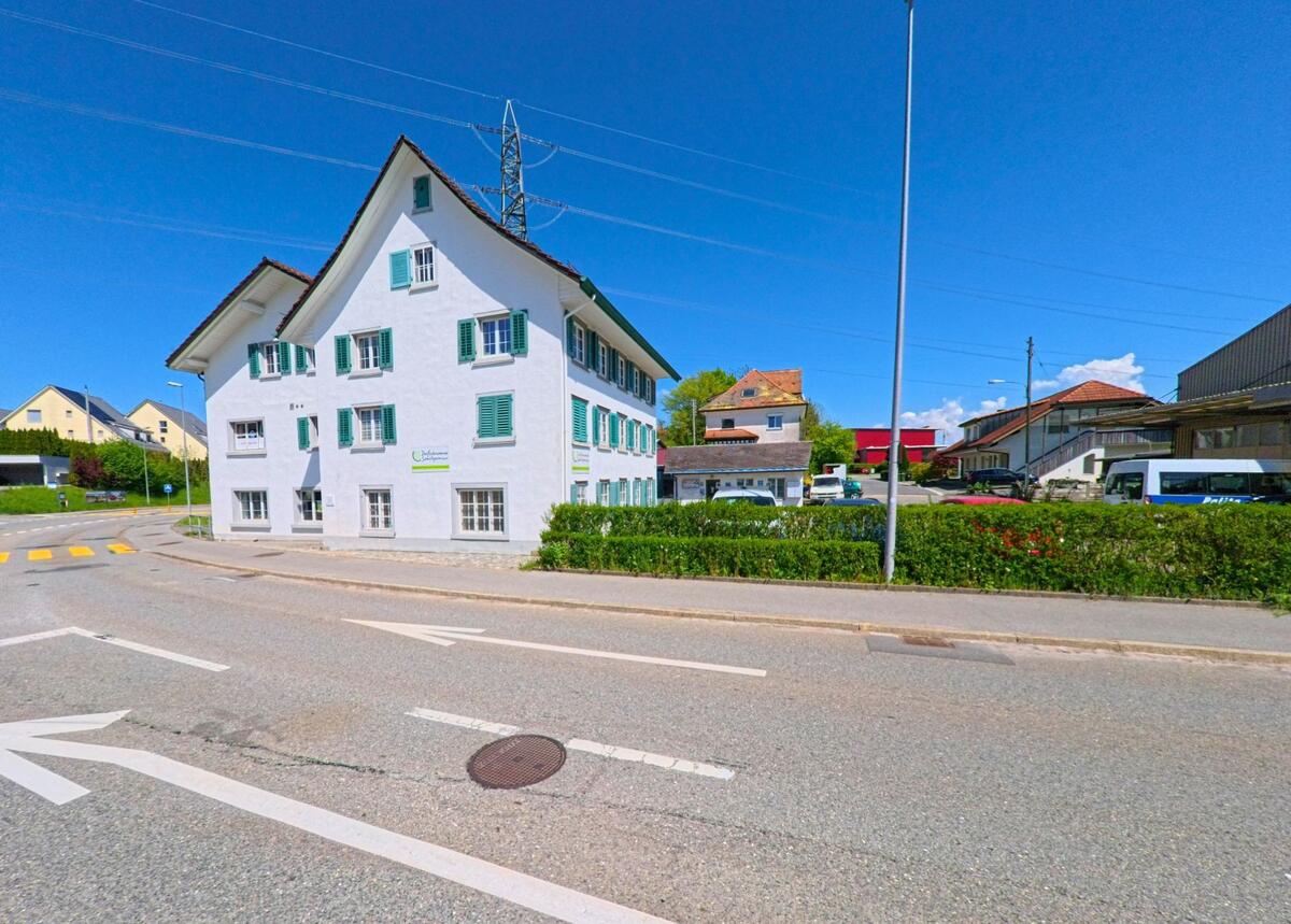 In Richterswil, 8833 im Kanton Zürich, steht ein weiss verputztes Eckhaus mit grünen Fensterläden an einer gut ausgebauten Kreuzung mit Gewerbebauten im Hintergrund.