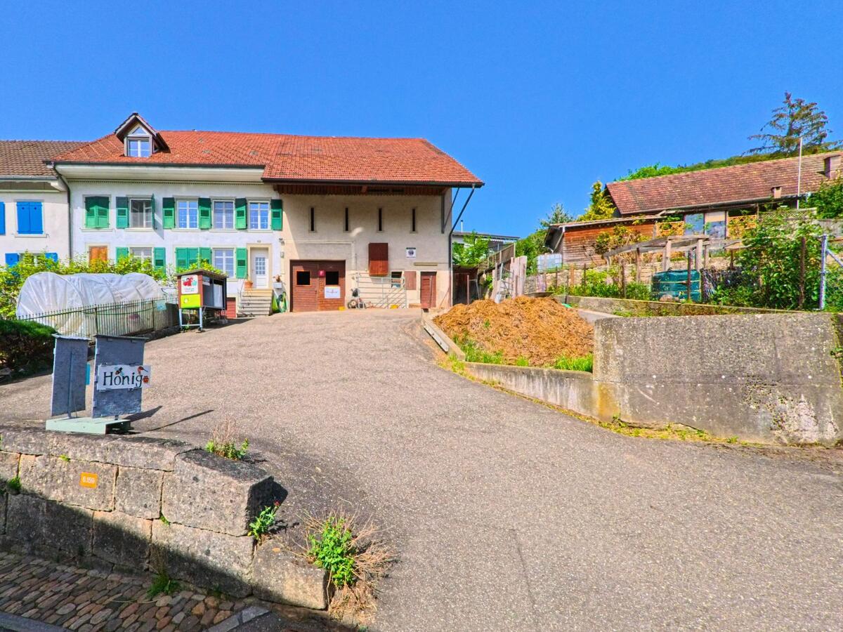 In Rickenbach (4462, Kanton Baselland) liegt ein Haus mit farbigen Fensterlaeden an einer steilen Zufahrt, daneben befinden sich ein Gewaechshaus und ein Hinweis auf die Ortsteilbezeichnung Hönnjo.