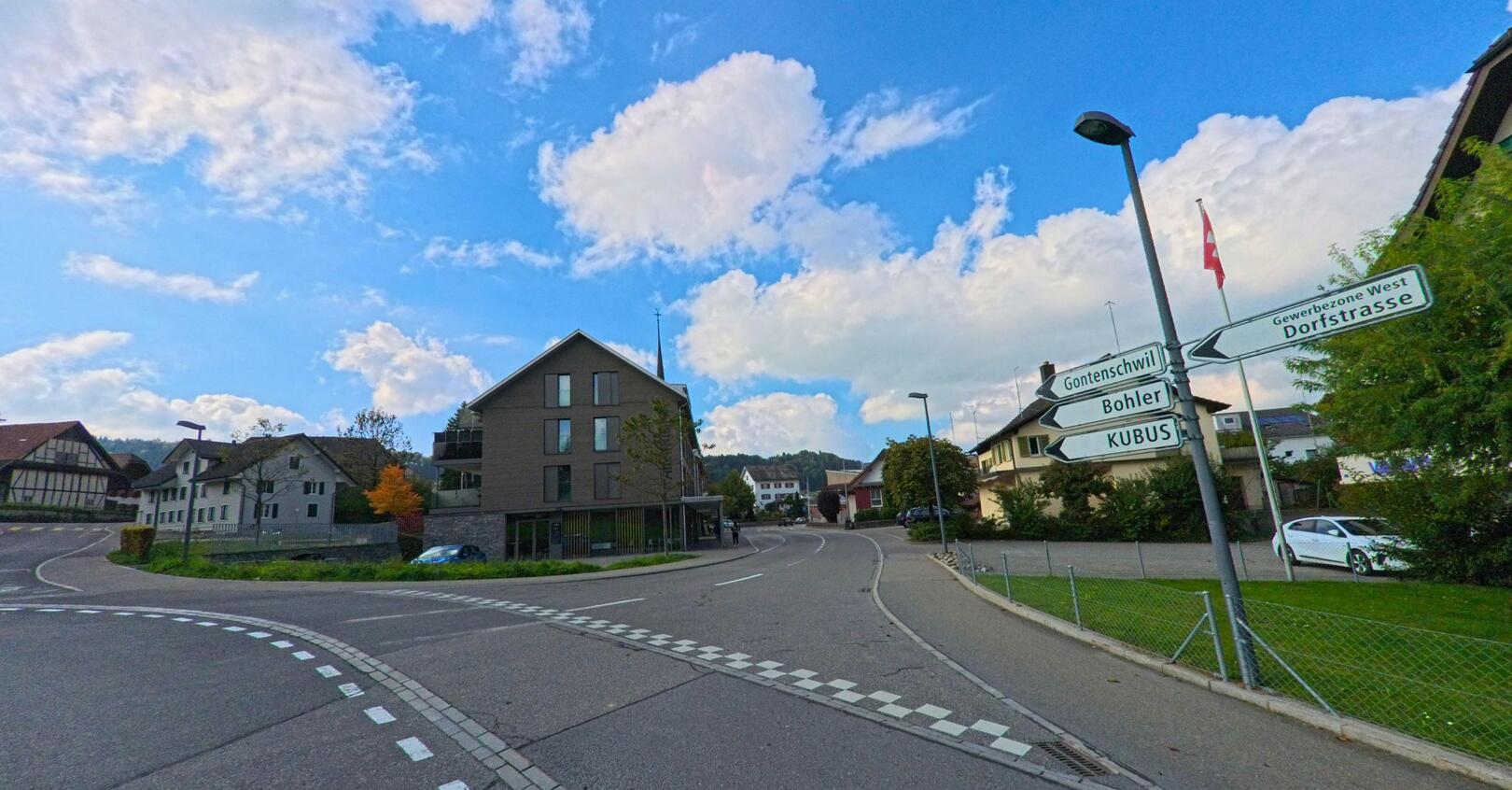 An einer leicht gekruemmten Strasse in Rickenbach (6221, Kanton Luzern) stehen Wohnhaeuser und Wegweiser, waehrend im Hintergrund Hügel und Wolken den Ort einrahmen.