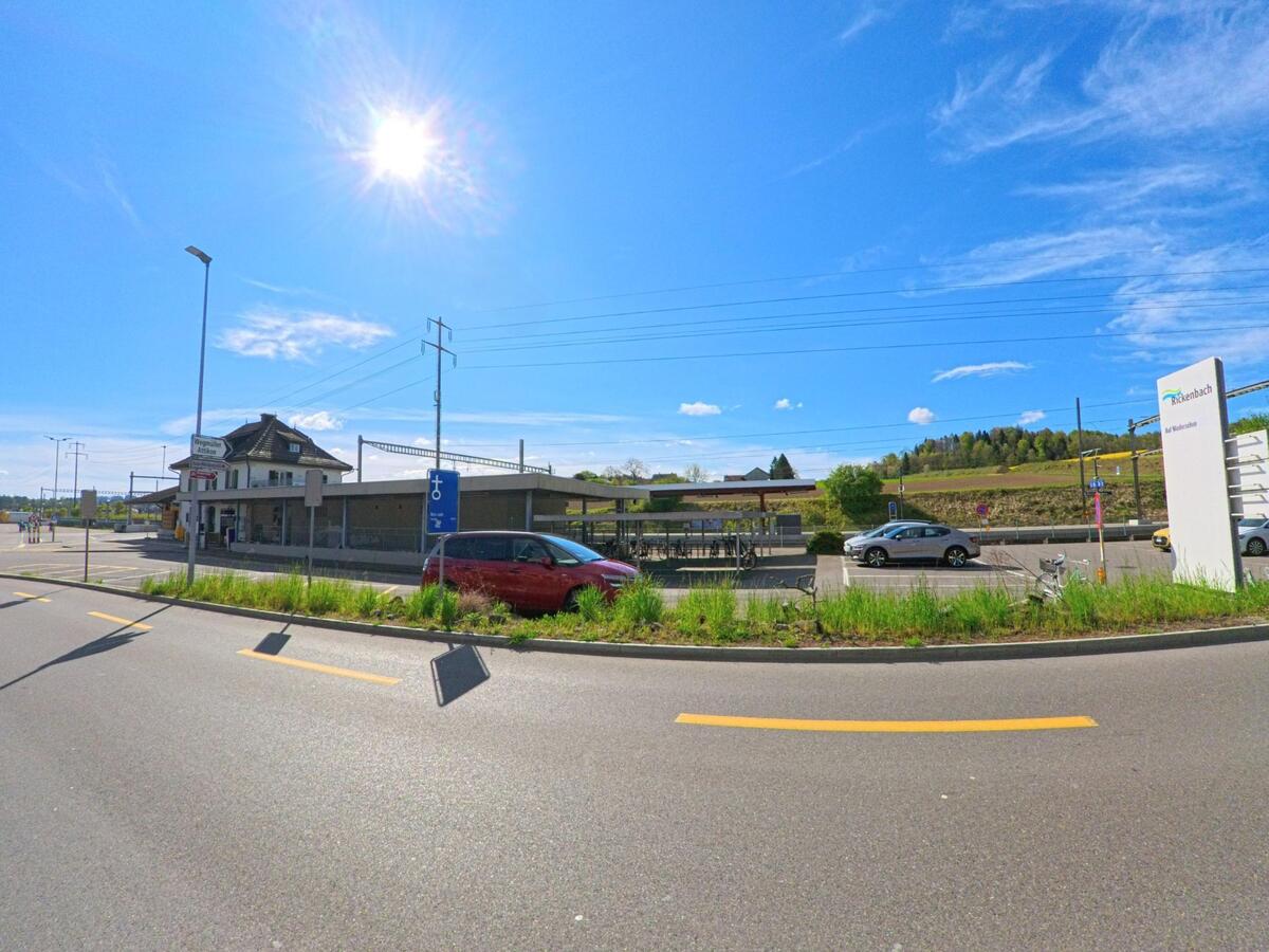Bei Sonnenschein liegt der Bahnhof von Rickenbach (8544, Kanton Zürich) mit angrenzendem Parkplatz und Blick auf die umliegenden Felder.