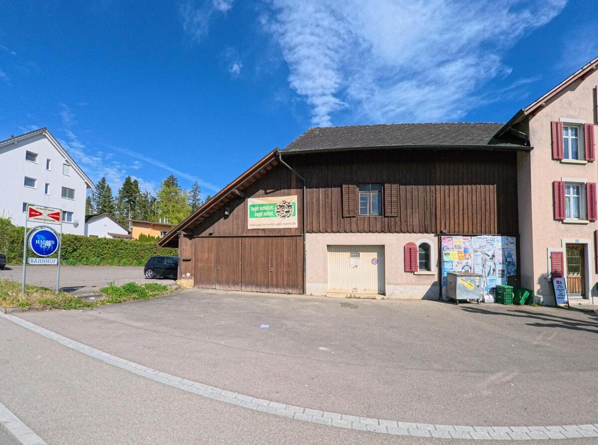 In Rickenbach (8544, Kanton Zürich) steht ein Gebäude mit Holzfassade und roten Fensterläden an einem Platz mit Anschlagtafel und Parkplätzen.