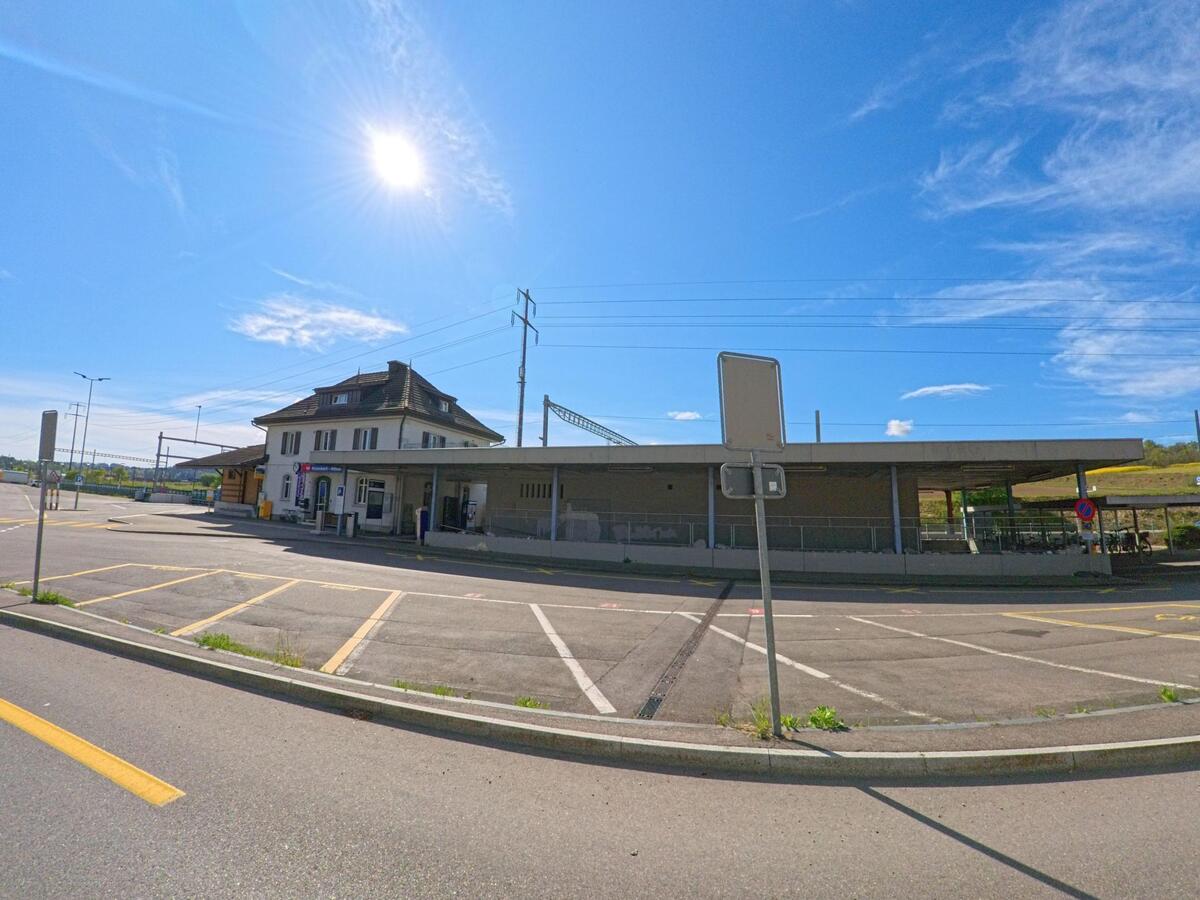 Unter blauem Himmel liegt in Rickenbach (8545, Kanton Zürich) ein Bahnhof mit Hauptgebäude, überdachtem Bahnsteig und angrenzendem Parkplatz.