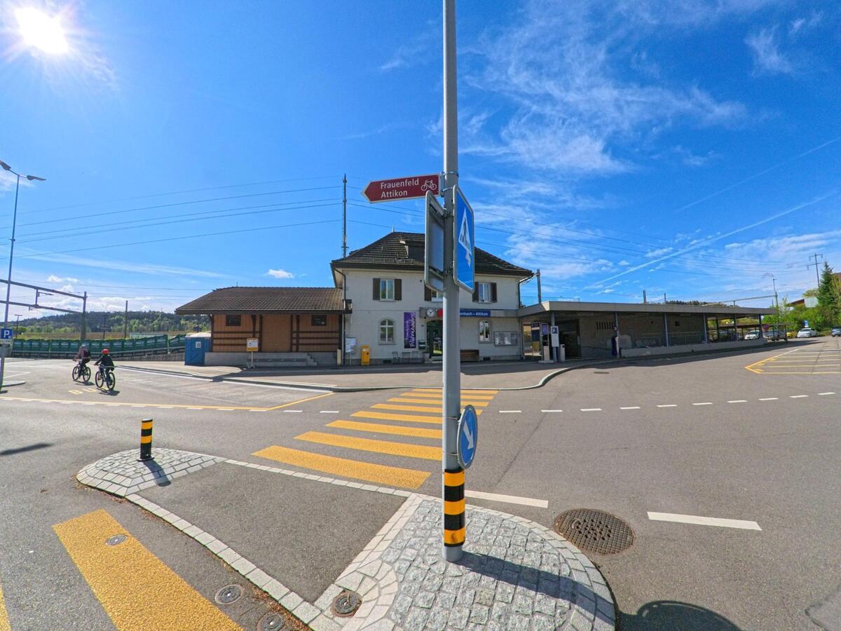 Vor dem Bahnhof in Rickenbach (8545, Kanton Zürich) verläuft ein Zebrastreifen, während zwei Radfahrer die Strasse bei sonnigem Wetter überqueren.