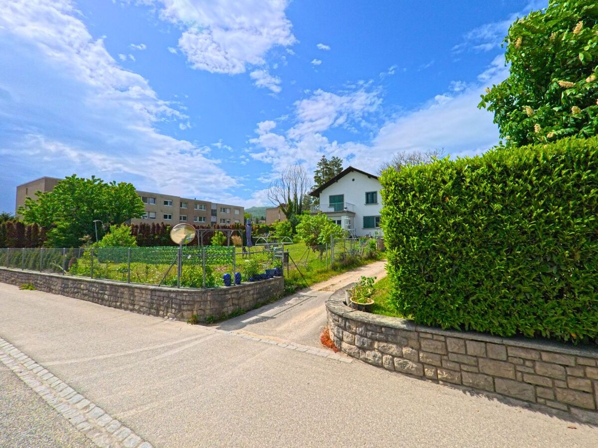 In Rickenbach (PLZ 4613, Kanton Solothurn) fuehrt ein schmaler Weg zwischen einem Wohnhaus und einem kleinen Garten mit Maschendrahtzaun vorbei.