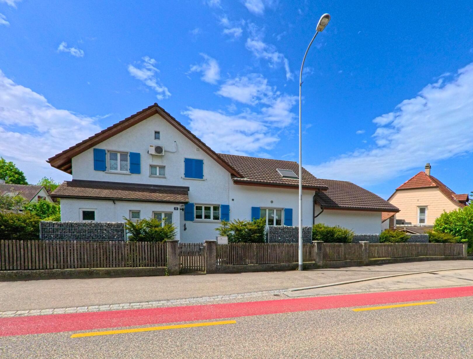 In Rickenbach (4613, Kanton Solothurn) stehen entlang einer Hauptstrasse mehrere Einfamilienhaeuser mit gepflegten Gaerten und farbigen Fensterlaeden.