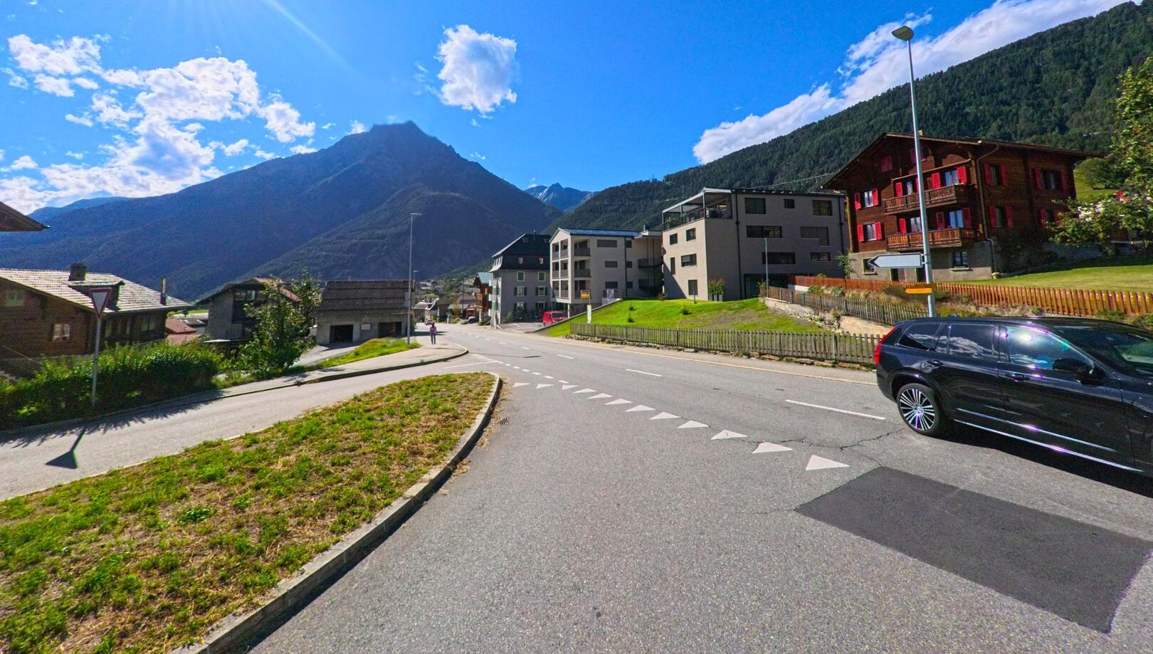 In Ried-Brig (3911, Kanton Wallis) verläuft eine Strasse mit moderner und traditioneller Bebauung, während im Hintergrund die umliegenden Berge unter blauem Himmel sichtbar sind.
