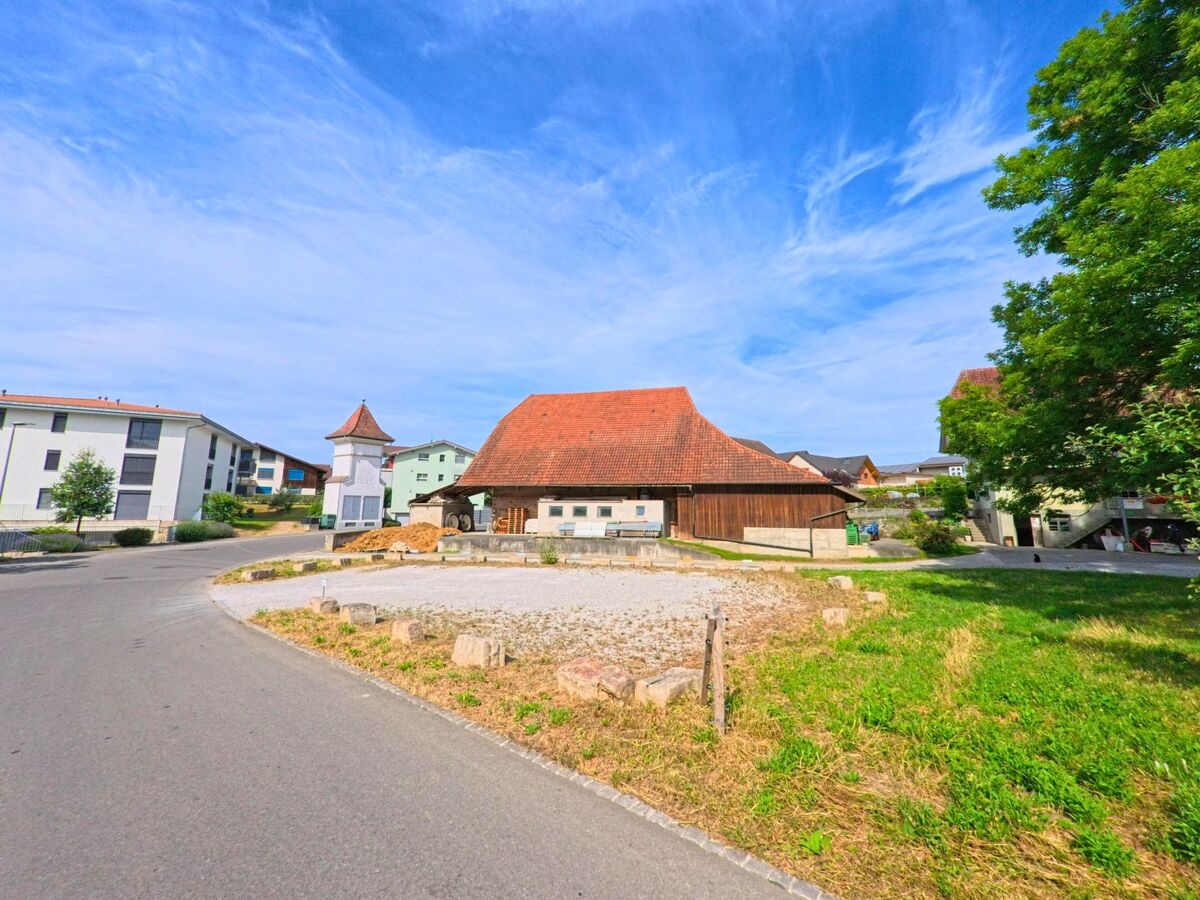 In Ried bei Kerzers (3216, Kanton Freiburg) steht ein traditioneller Bauernhof mit rotem Ziegeldach neben einem kleinen weissen Turm im Kontrast zu modernen Wohnbauten.
