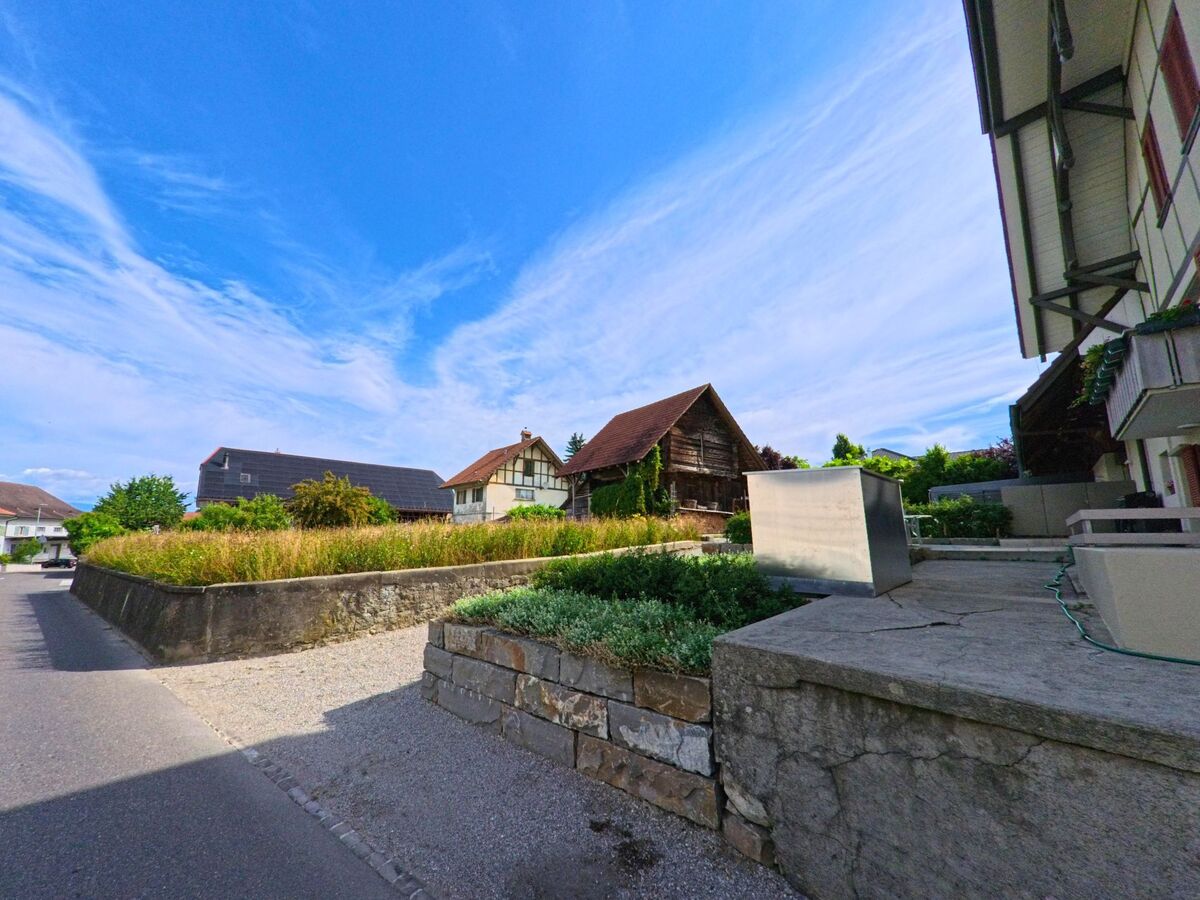 In Ried bei Kerzers (3216, Kanton Freiburg) verläuft eine schmale Quartierstrasse entlang gepflegter Gärten und traditioneller Wohnhäuser mit teils hölzerner Fassade.