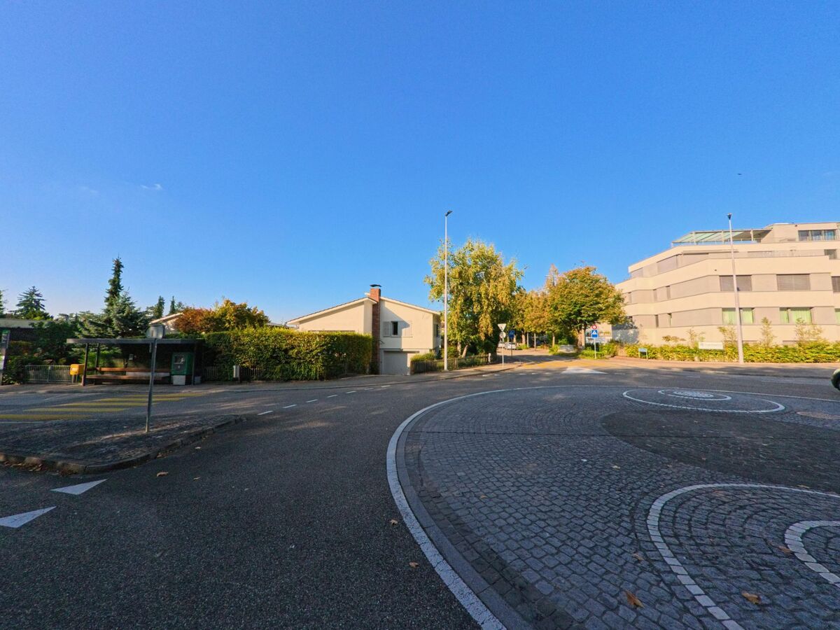 An einem kleinen Kreisel in Riehen (4125, Kanton Basel-Stadt) treffen moderne Wohnbauten, eine Bushaltestelle und begruente Hauser bei klarem Himmel aufeinander.
