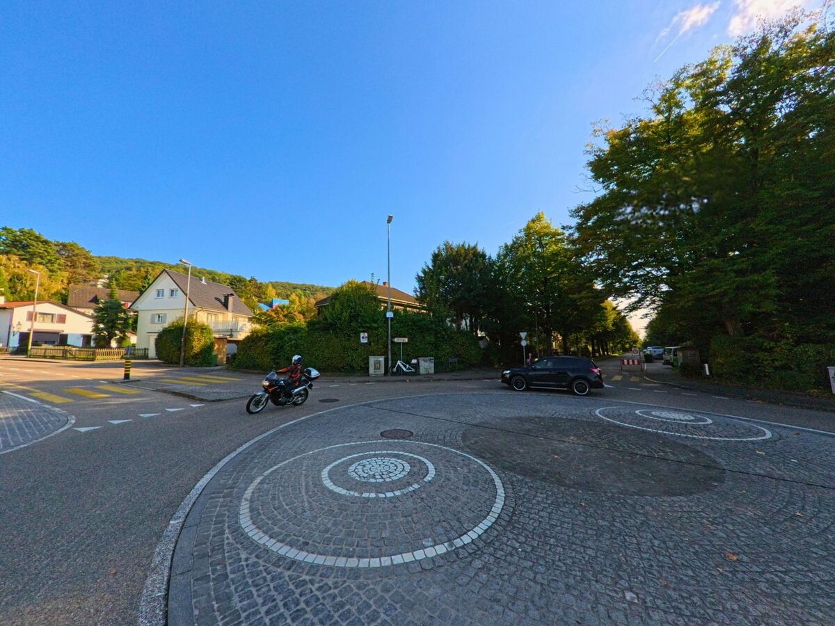 Ein Kreisel in Riehen (4125, Kanton Basel-Stadt) verbindet eine von Baeumen gesaeumte Strasse mit einem Wohnquartier, waehrend ein Motorradfahrer vorbeifaehrt.