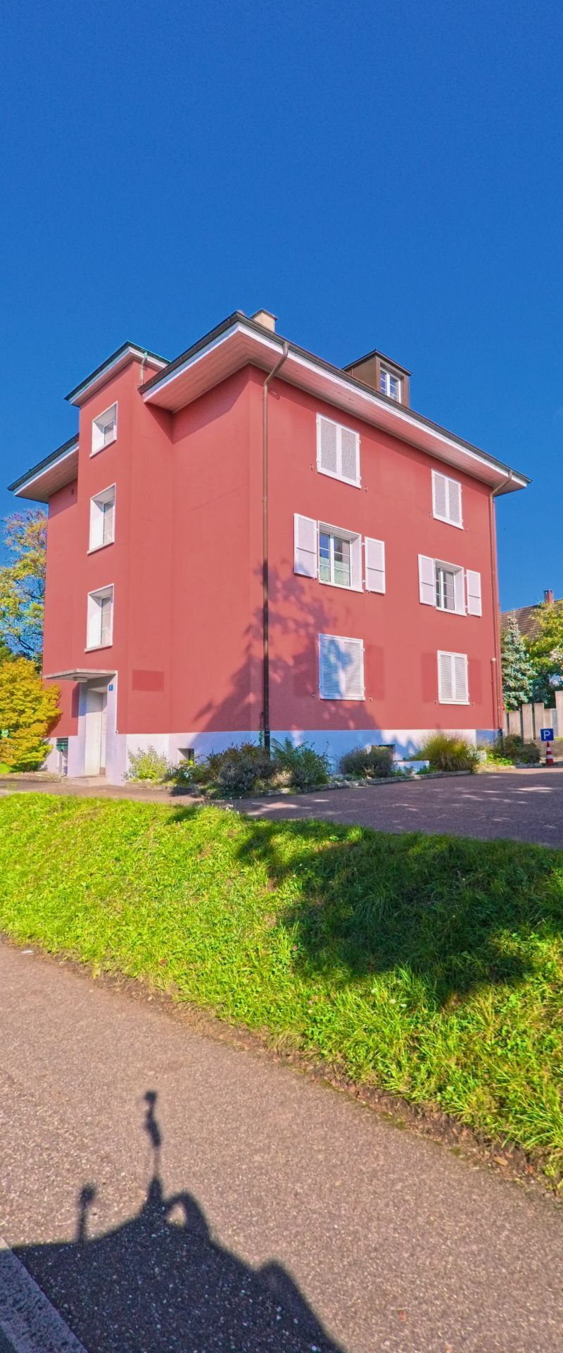 Ein rosafarbenes Mehrfamilienhaus mit weissen Fensterlaeden steht in Riehen (4125, Kanton Basel-Stadt) am Rand eines sonnigen Gehwegs mit niedriger Begruenung.