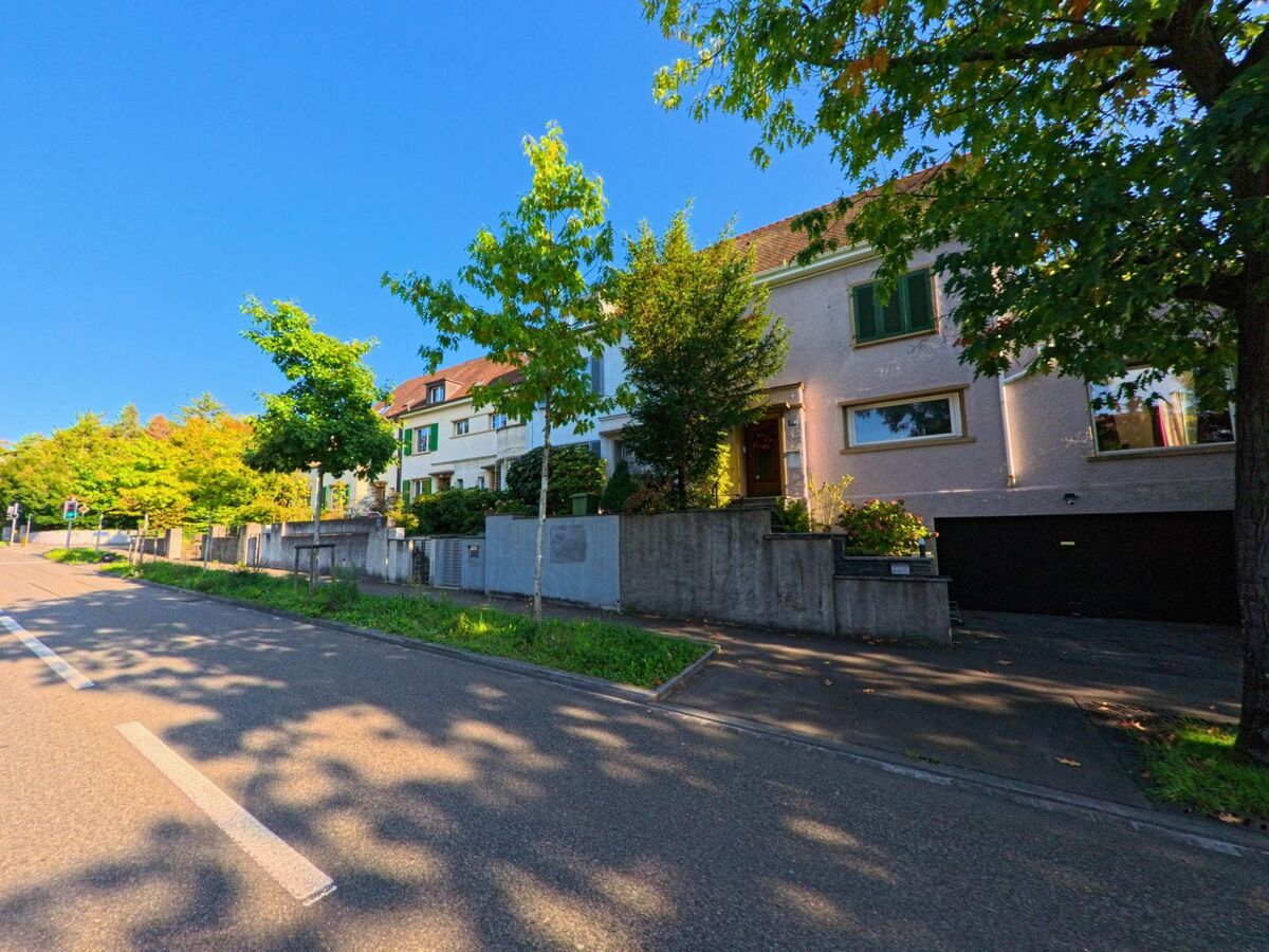 Entlang einer ruhigen Strasse in Riehen (4125, Kanton Basel-Stadt) stehen gepflegte Wohnhaeuser hinter niedrigen Mauern und jungen Baemen im Sonnenlicht.