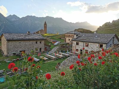 Alt-Text: In Brusio (7743, Kanton Graubuenden) liegen Steinhaeuser mit einer kleinen Kirche auf einer Bergwiese, umgeben von roten Blumen und hohen Gipfeln.