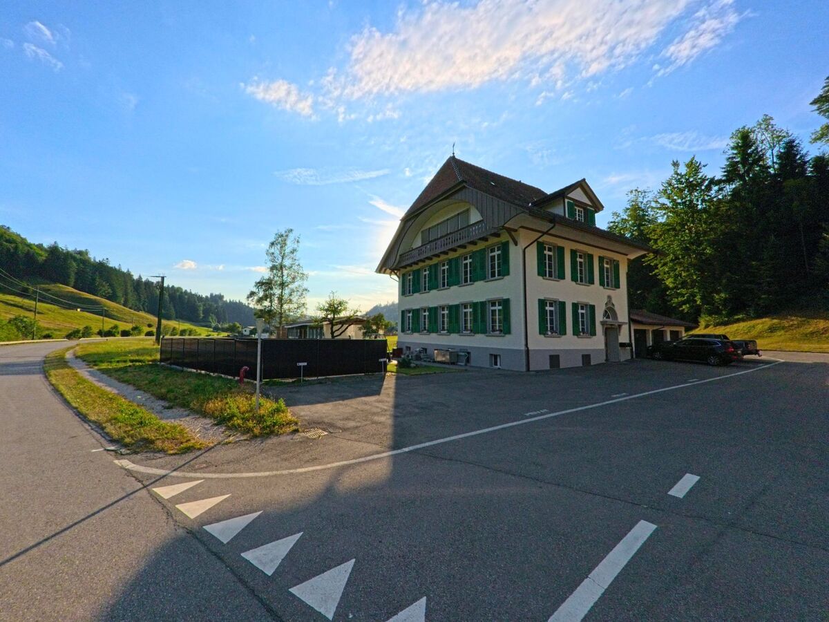 Am Waldrand von Riggisberg (3099, Kanton Bern) steht ein dreigeschossiges Gebäude mit grünen Fensterläden und symmetrischer Fassade neben einer Zufahrtsstrasse.
