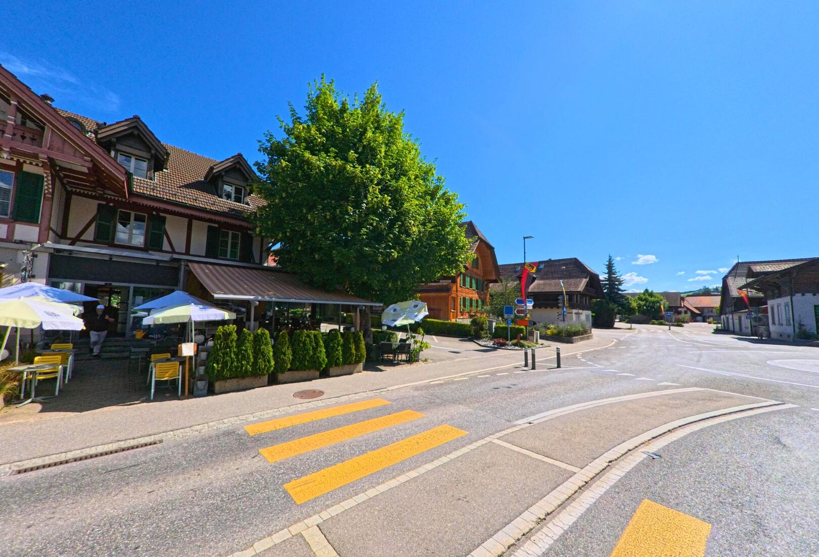 In Riggisberg (3132, Kanton Bern) lädt ein Café mit Sonnenschirmen an einer ruhigen Kreuzung mit traditionellen Häusern zum Verweilen ein.