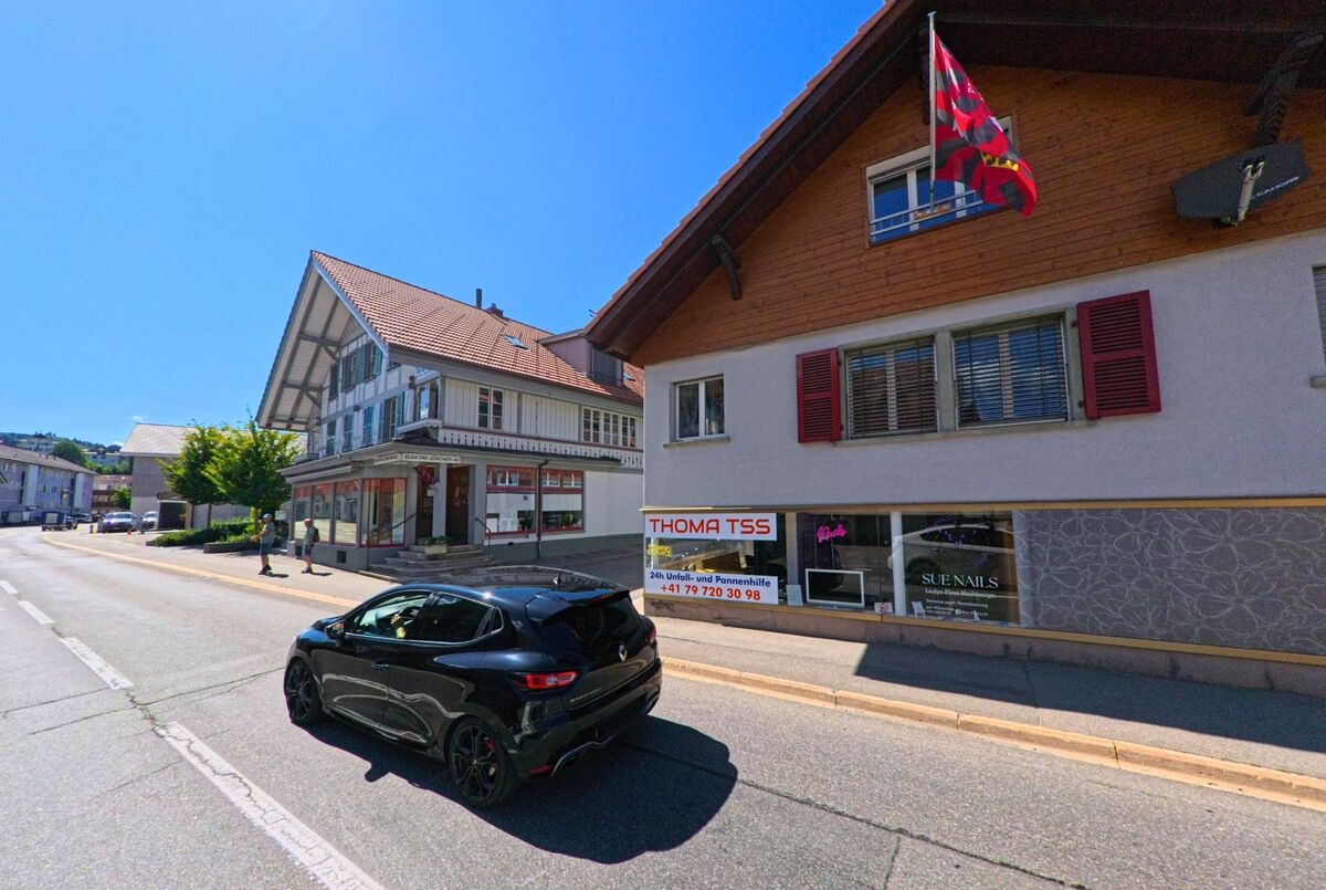 Entlang der Hauptstrasse von Riggisberg (3132, Kanton Bern) reihen sich traditionelle und moderne Gebäude, darunter ein Nagelstudio und ein Pannendienst.
