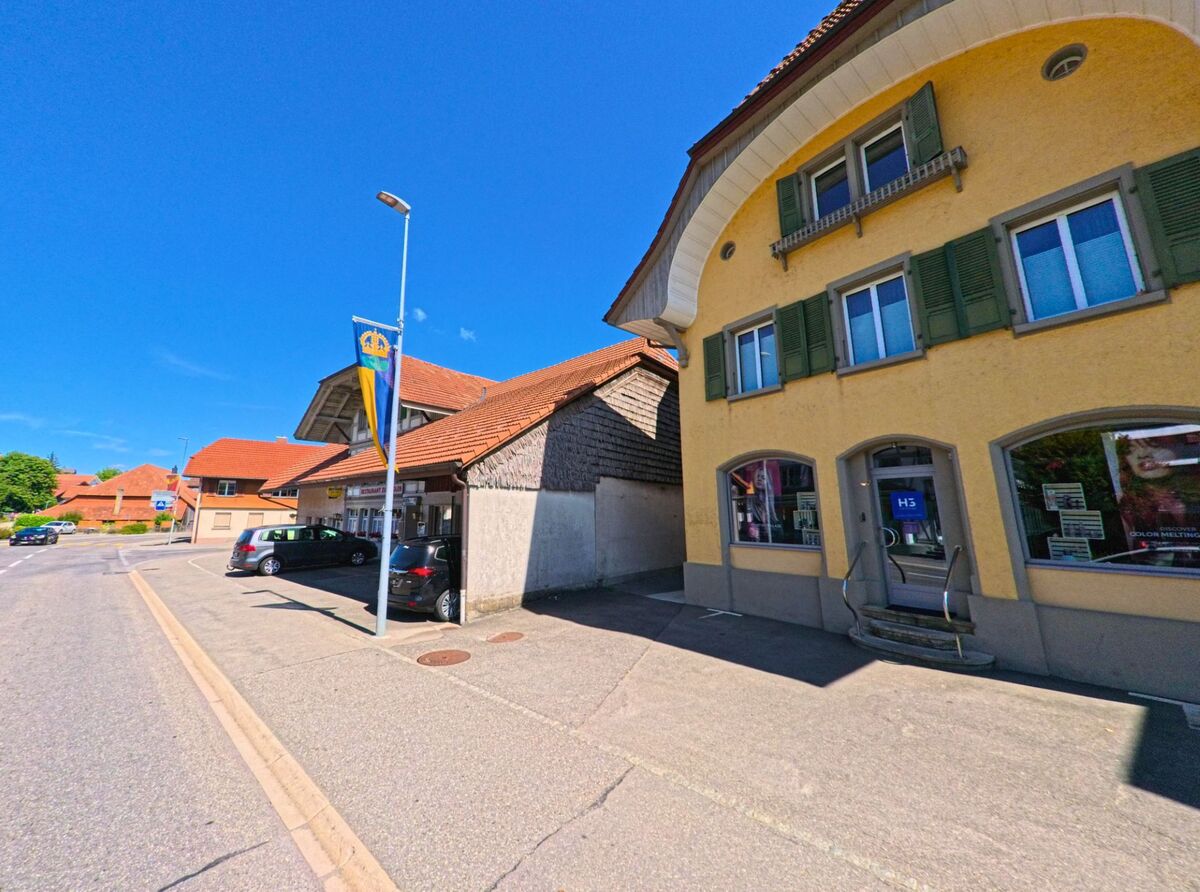 In Riggisberg (3132, Kanton Bern) stehen entlang einer Nebenstrasse verschiedene Geschäfte in traditionellen Gebäuden mit roten Dächern und bunten Fassaden.
