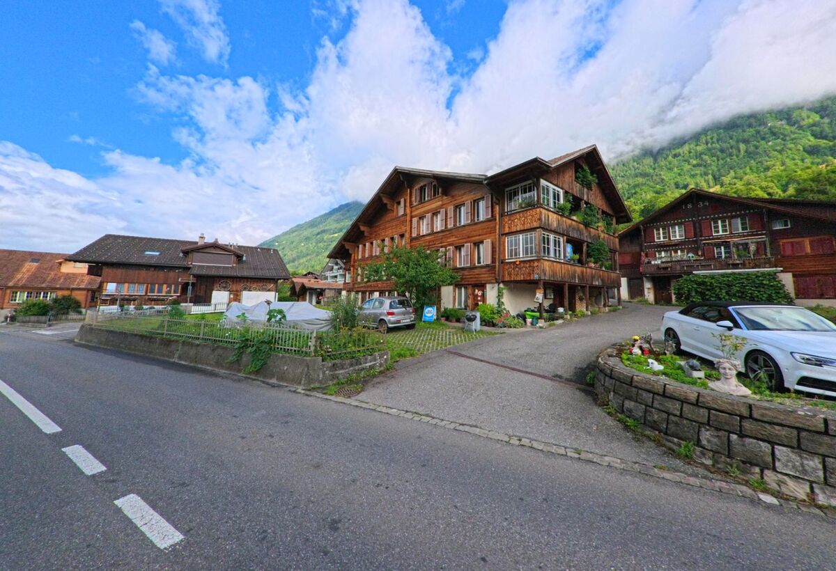 In Ringgenberg (3852, Kanton Bern) stehen mehrere traditionelle Holzhäuser mit Blumenbalkonen an einer ruhigen Nebenstrasse vor einem bewaldeten Berghang.