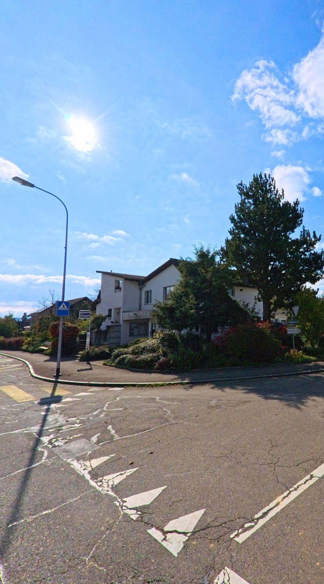 An einer Kreuzung in Riniken (5223, Kanton Aargau) steht ein Wohnhaus mit grossen Baeumen und gepflegter Bepflanzung bei strahlendem Sonnenschein.