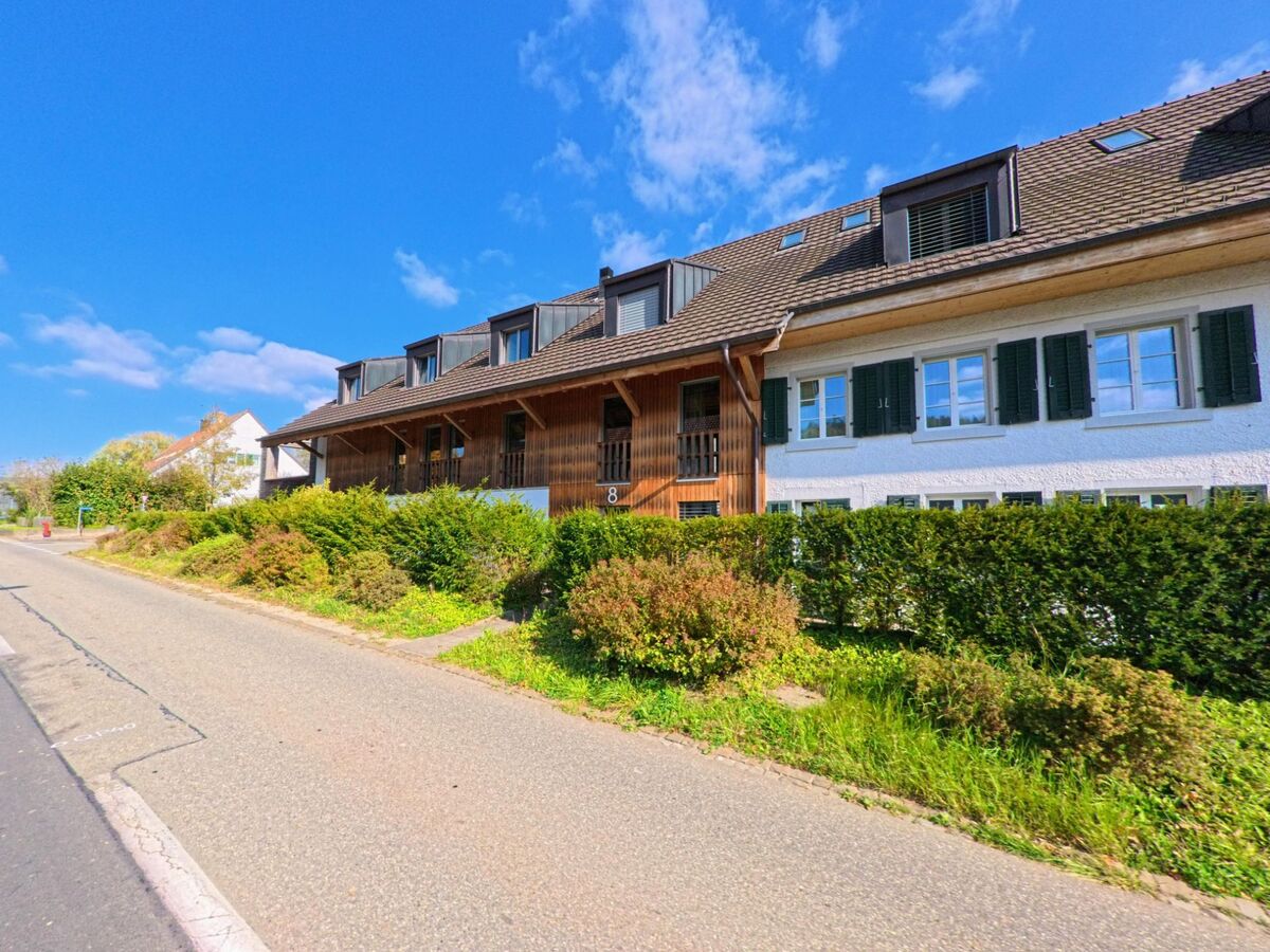 Ein laengliches Wohnhaus mit Holzfassade und gruenen Fensterlaeden steht an einer ruhigen Strasse in Riniken (5223, Kanton Aargau).