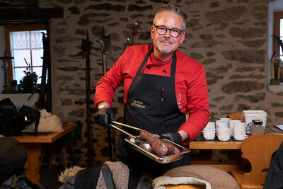 Im Ristorante Molinari in Simplon (3907, Kanton Wallis) präsentiert ein Koch in roter Jacke und schwarzer Schürze eine frisch zubereitete Fleischspezialität in einem rustikalen Gastraum mit Steinwänden.