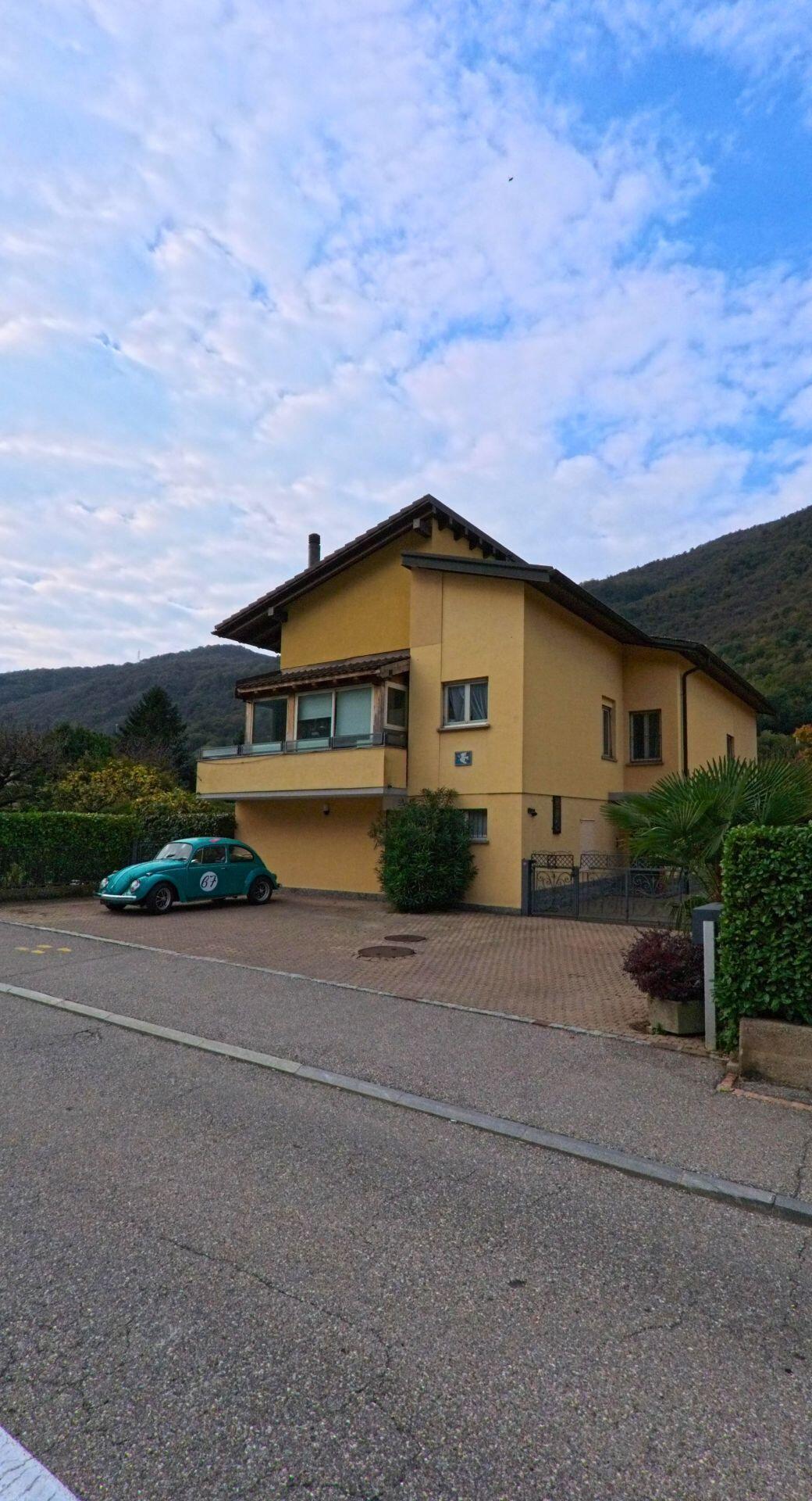 Ein gelbes Wohnhaus in Riva San Vitale (6826, Kanton Tessin) steht am Hang unter blauem Himmel, vor dem Haus parkt ein alter VW Kafer.