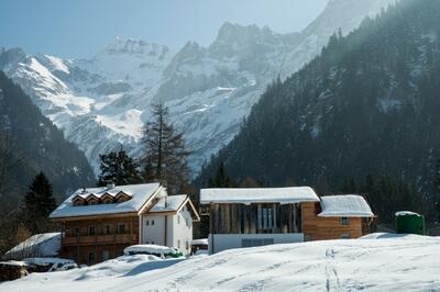 In Les Plans-sur-Bex (1880, Kanton Waadt) stehen zwei Chalets mit verschneiten Dächern vor einer imposanten Bergkulisse aus Felsen und Tannen.