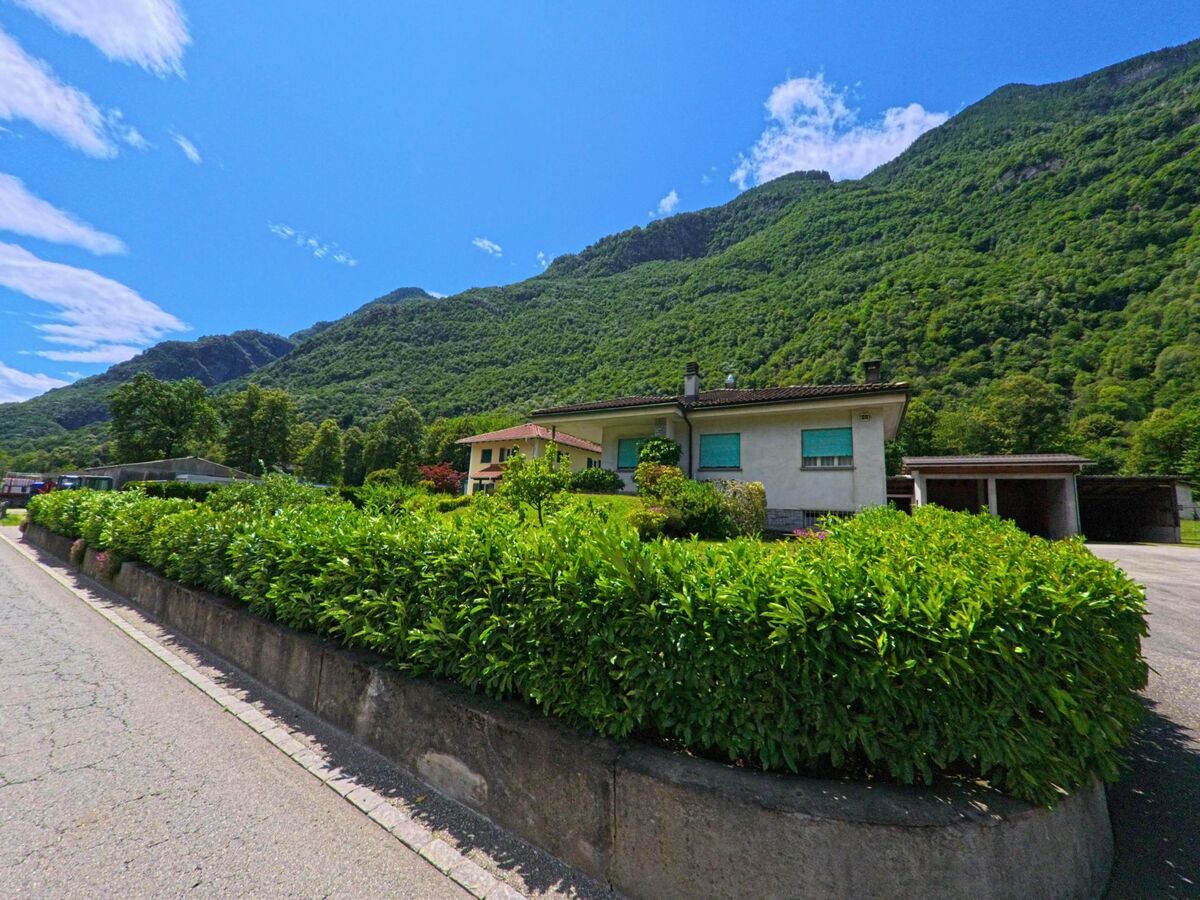Ein Einfamilienhaus mit gepflegter Hecke und Garage steht in Riviera (6526, Kanton Tessin) vor einer steilen, dicht bewaldeten Bergflanke.