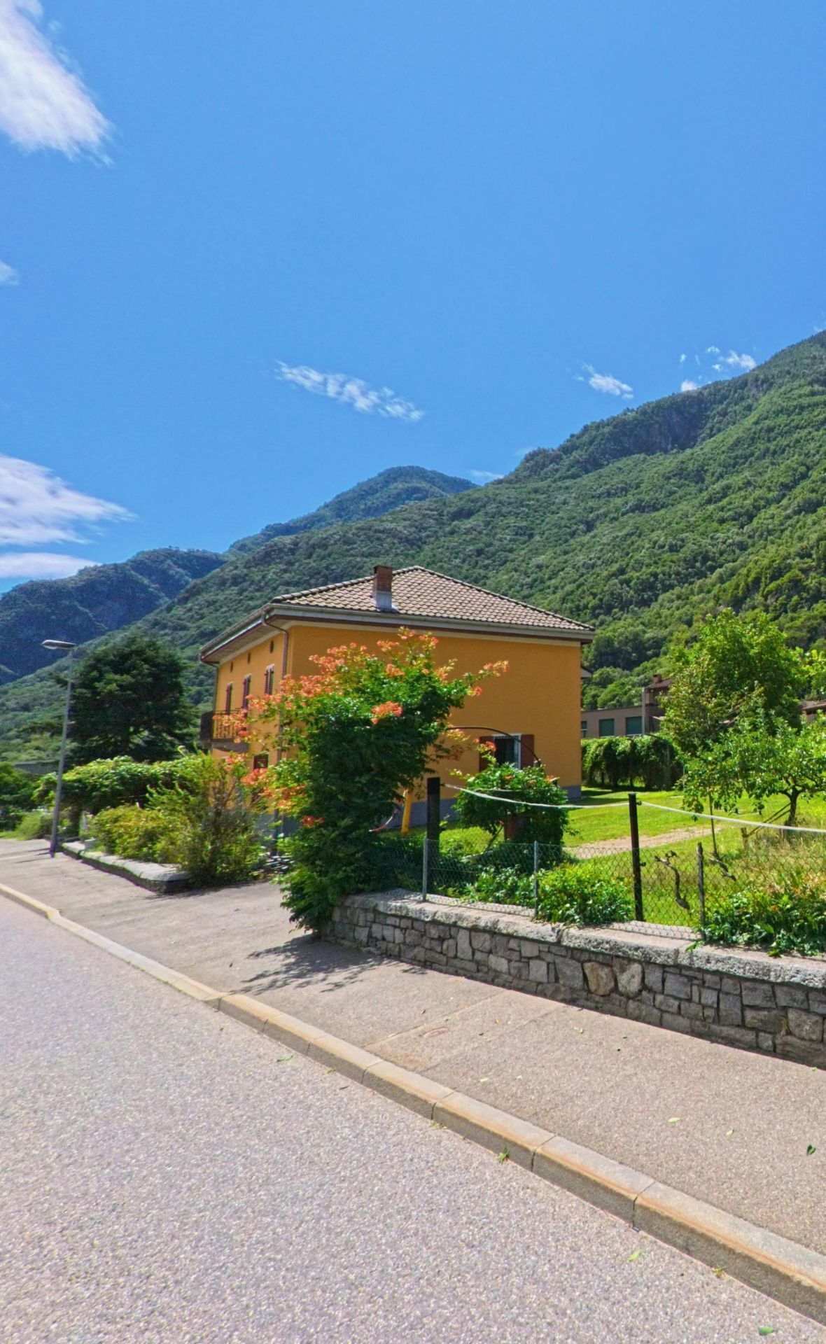 Ein gelbes Wohnhaus mit Garten steht in Riviera (6526, Kanton Tessin) am Strassenrand vor der Kulisse der grünen Berge.
