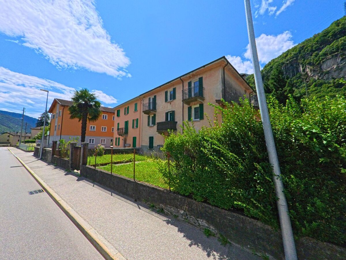 Mehrfamilienhäuser mit farbigen Fassaden und grünen Fensterläden stehen in Riviera (6527, Kanton Tessin) an einer sonnigen Strasse mit Blick auf bewaldete Hänge.