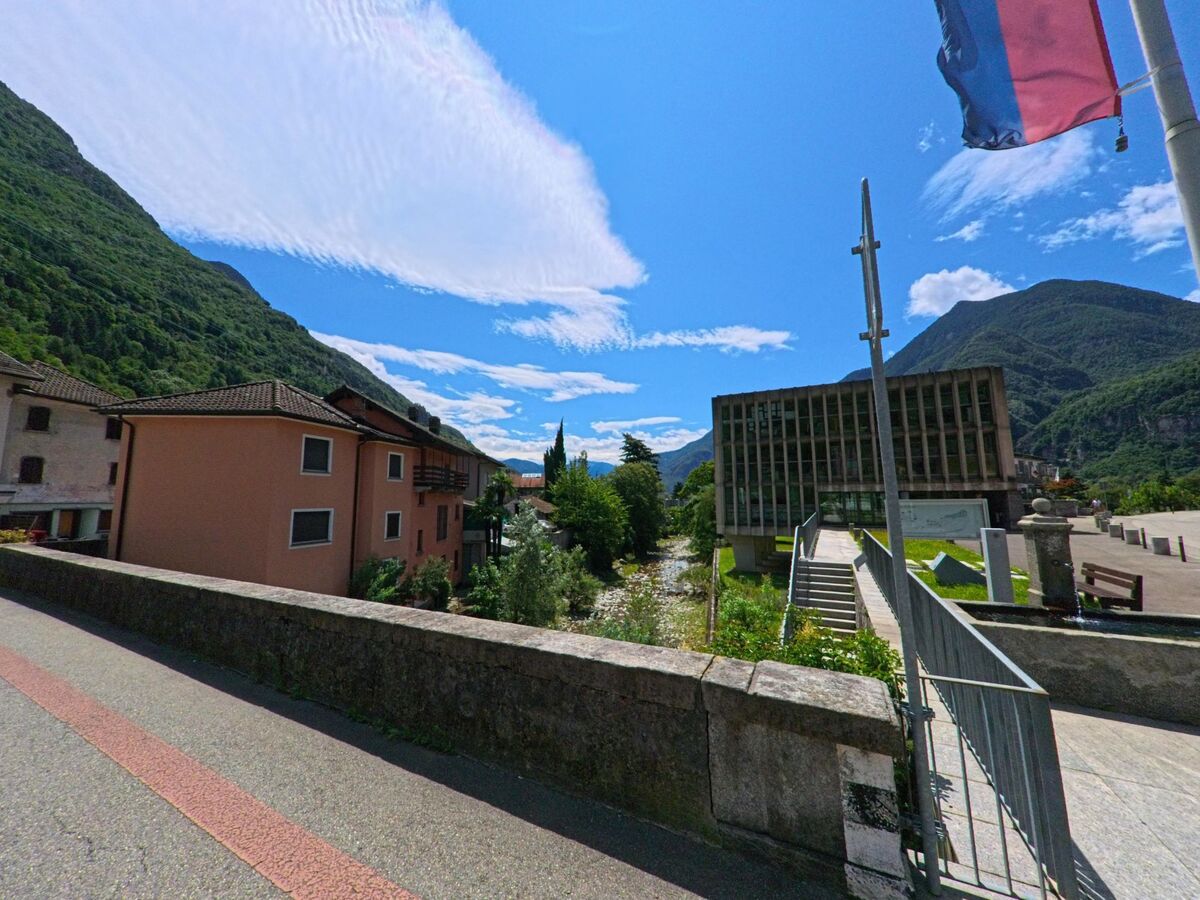 Über eine Brücke in Riviera (6703, Kanton Tessin) blickt man auf den Flusslauf zwischen modernen und traditionellen Gebäuden, umgeben von bewaldeten Bergen.