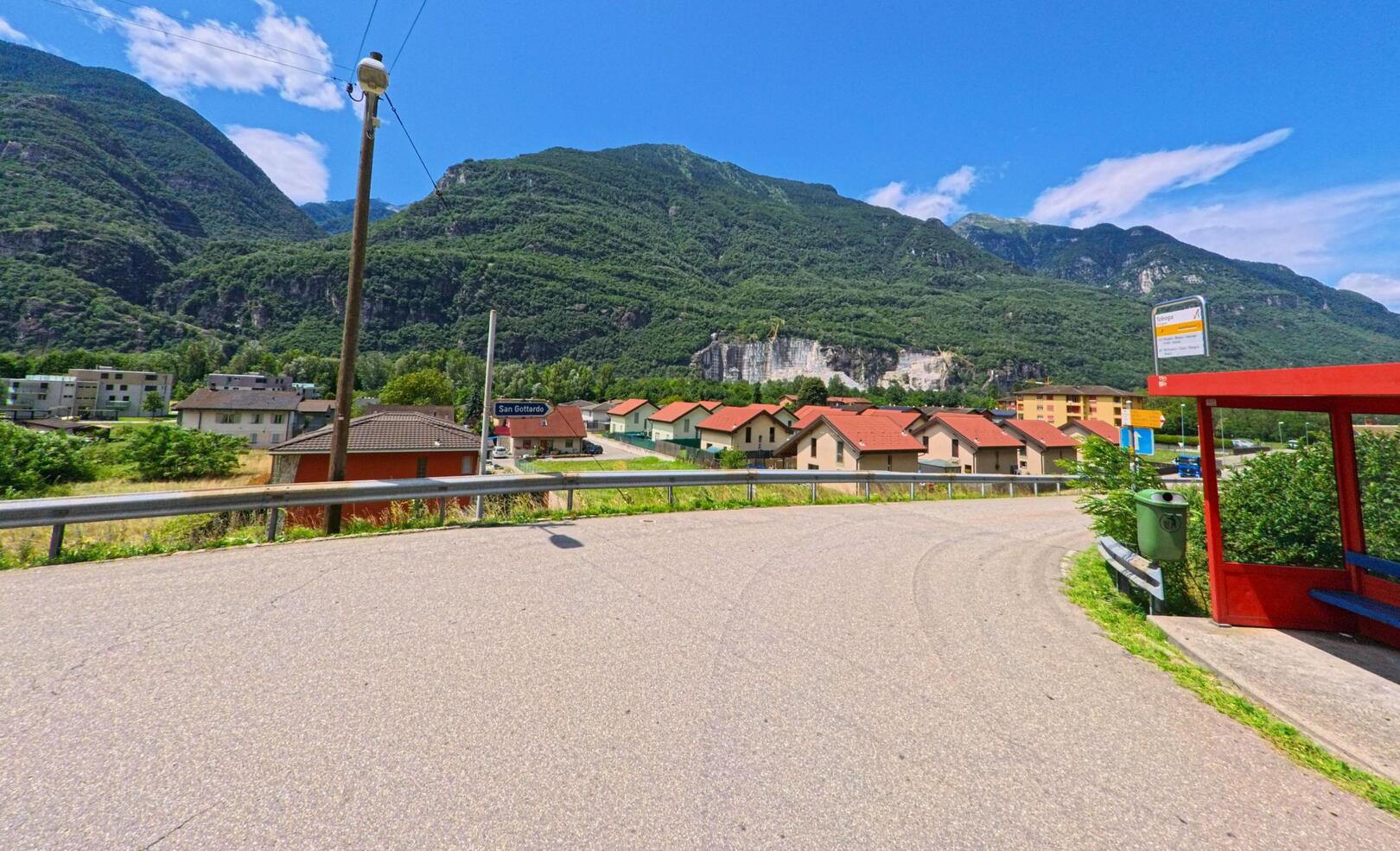 An einer Haltestelle in Riviera (6703, Kanton Tessin) bietet sich ein weiter Blick über Wohnhäuser mit roten Dächern und die bewaldeten Hänge der umliegenden Berge.