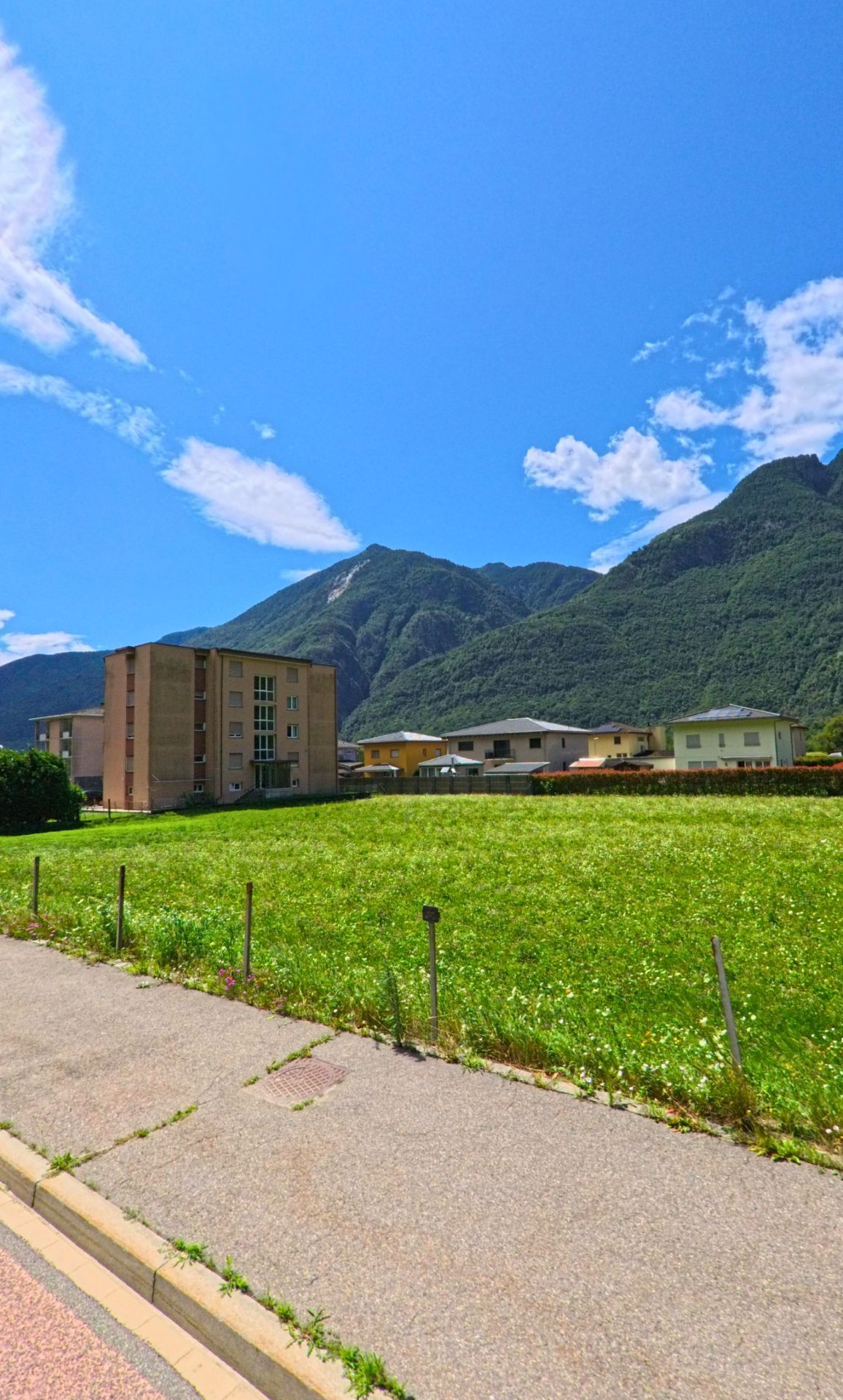 Eine Strasse in Riviera (6705, Kanton Tessin) führt vorbei am rot gestrichenen Gemeindehaus und an wehenden Fahnen mit Blick auf die umliegenden Berge.
