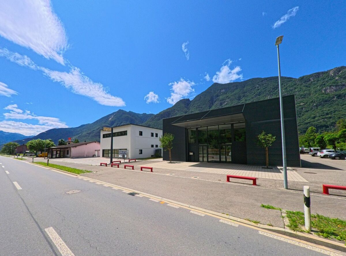 Entlang der Hauptstrasse von Riviera (6705, Kanton Tessin) stehen moderne Gewerbegebäude mit grossen Glasfronten vor der Kulisse der grünen Berglandschaft.