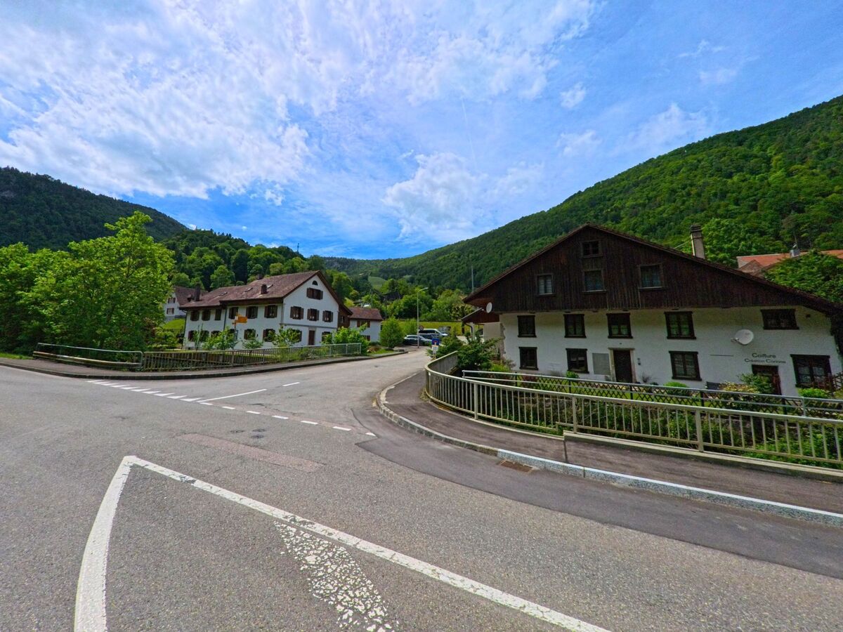 Im Zentrum von Roches (2762, Kanton Bern) zweigt eine Strasse zwischen traditionellen Wohnhäusern mit dunklem Holz und weissen Fassaden ab, eingebettet in eine grüne Hügellandschaft.