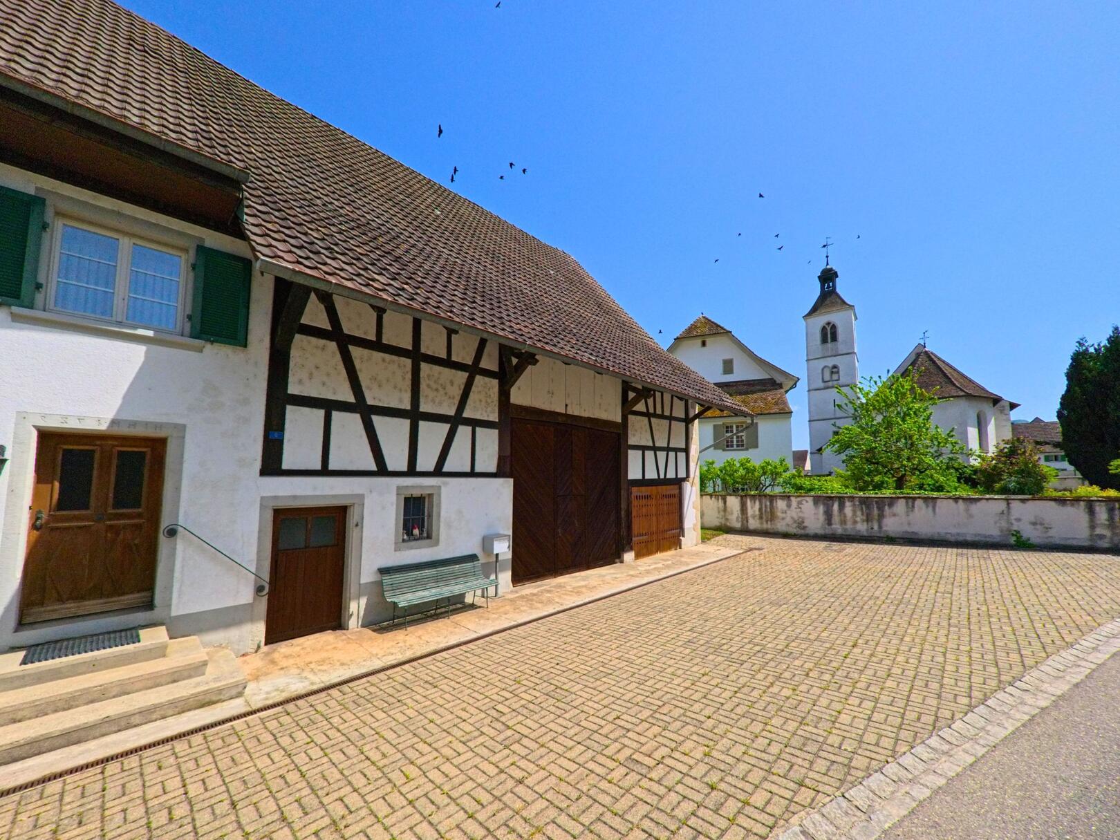 In Rodersdorf (4118, Kanton Solothurn) ist ein traditionelles Fachwerkhaus zu sehen, daneben ragt der Kirchturm der örtlichen Kirche in den blauen Himmel.