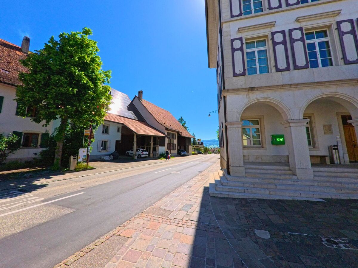 In Röschenz (4244, Kanton Baselland) führt eine Strasse an einem historischen Gebäude mit Arkaden und grünen Bäumen entlang, bei strahlendem Sonnenschein.