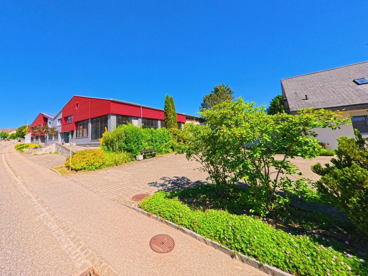 In Röschenz (4244, Kanton Baselland) führt eine Strasse an modernen, rot gestrichenen Gebäuden und gepflegten Grünflächen vorbei, unter einem klaren blauen Himmel.