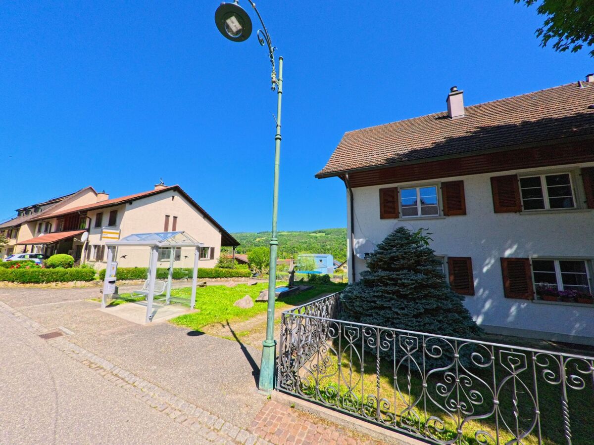 In Roggenburg (2814, Kanton Baselland) führt eine Strasse an einem Gebäude mit einem Buswartehäuschen und einem Laternenmast entlang, mit grünen Hügeln im Hintergrund.