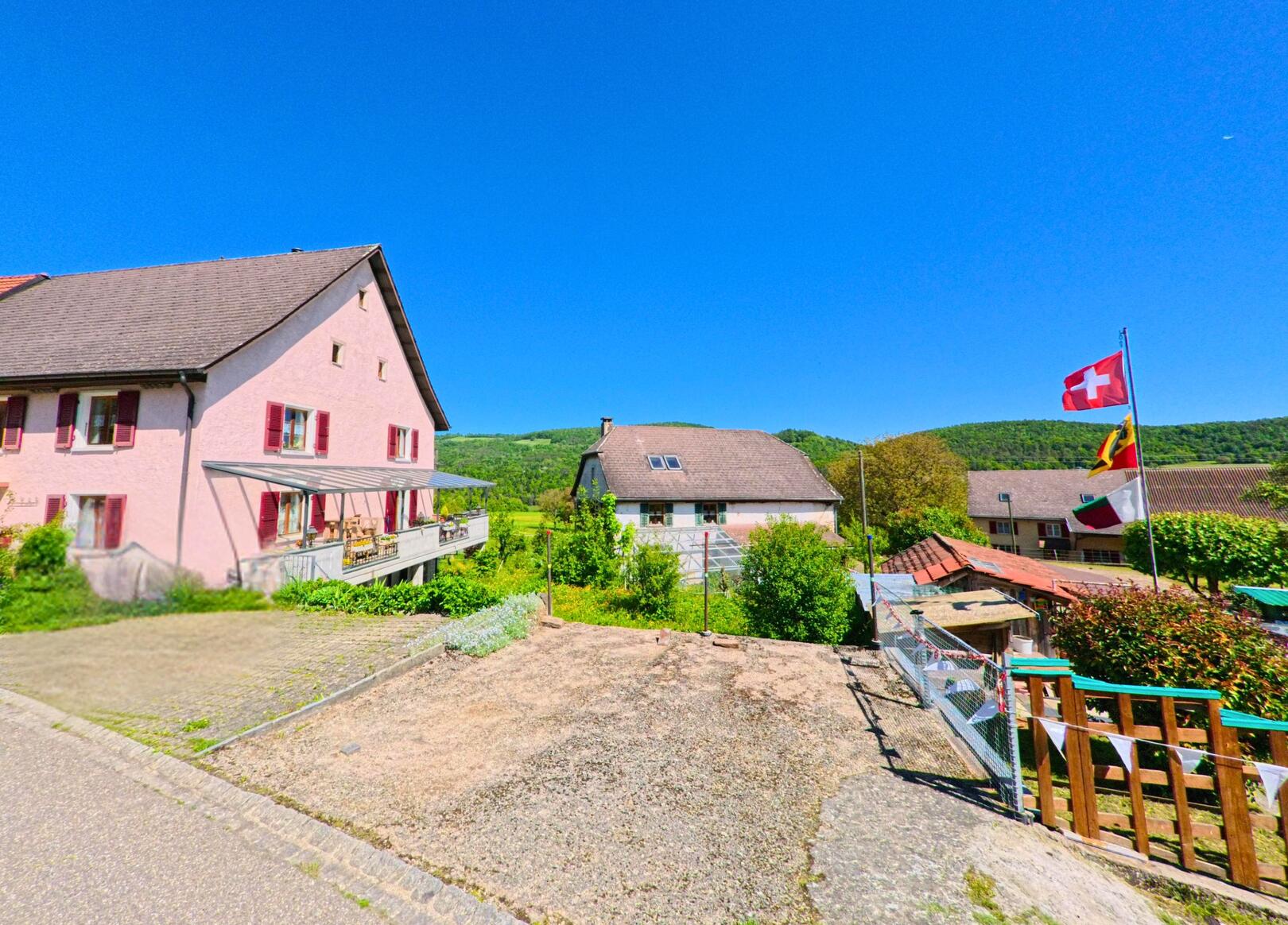 In Roggenburg (2814, Kanton Baselland) führt eine Strasse an einem pinkfarbenen Gebäude mit einem Auto und mehreren Flaggen, darunter die Schweizer Fahne, vorbei, mit grünen Hügeln im Hintergrund.
