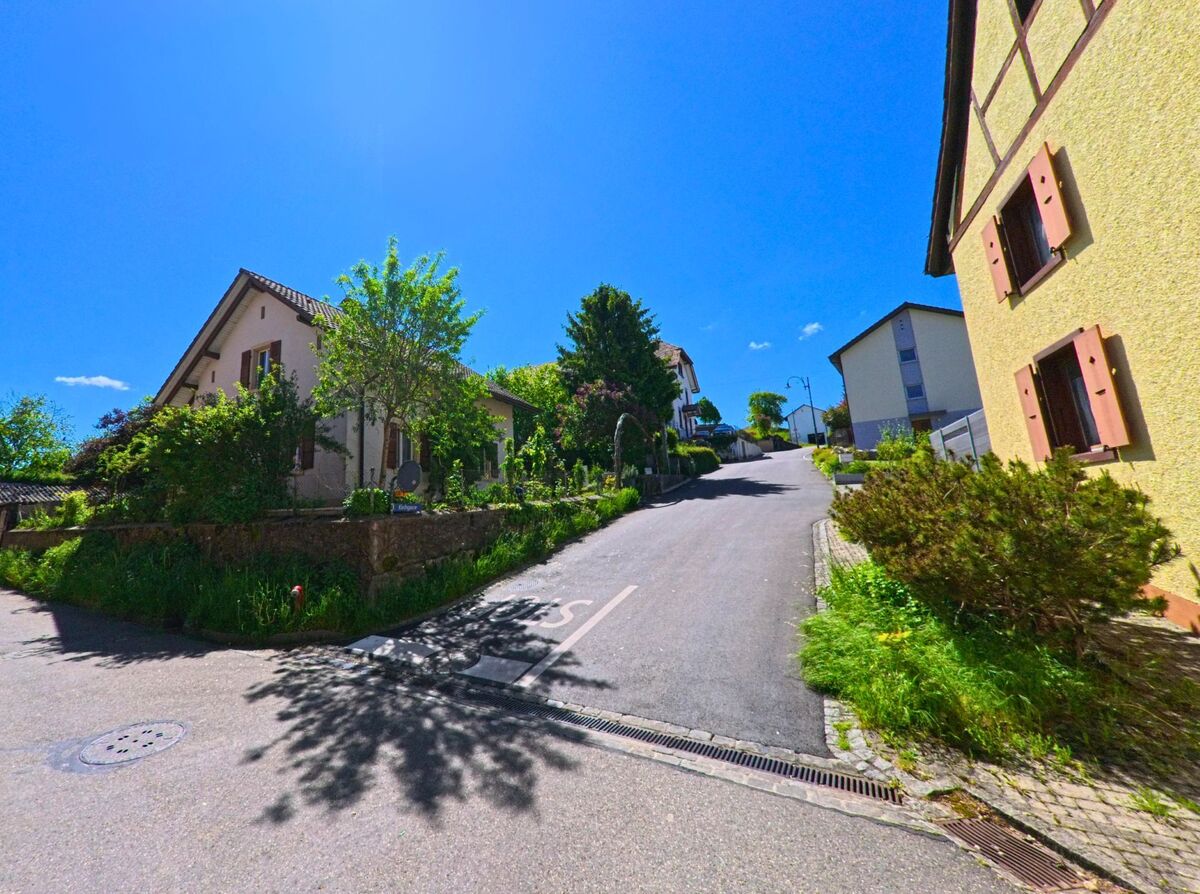 In Roggenburg (2814, Kanton Baselland) führt eine Strasse an einem charmanten Gebäude mit Garten entlang, mit einem sonnigen Himmel und grünen Hügeln im Hintergrund.