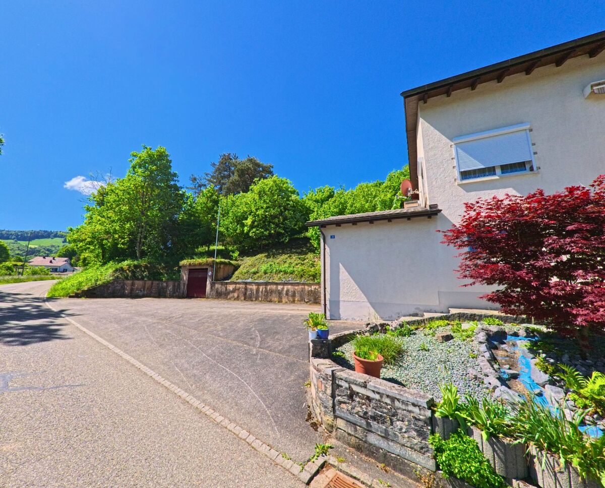 In Roggenburg (2814, Kanton Baselland) führt eine Strasse an einem Haus mit einem gepflegten Garten und einem roten Tor vorbei, während im Hintergrund grüne Hügel sichtbar sind.