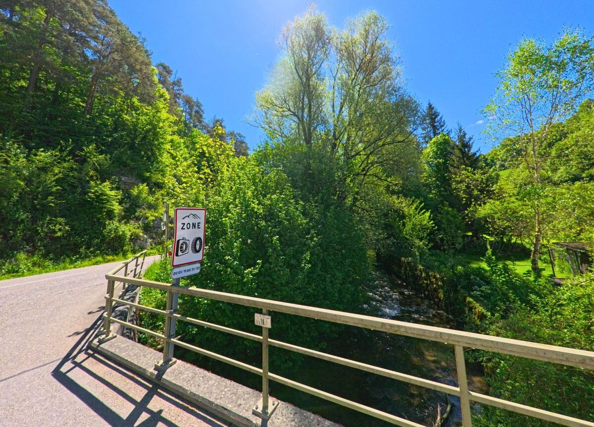 In Roggenburg (2814, Kanton Baselland) zeigt sich eine Strasse, die über eine Brücke führt, mit einem Verkehrsschild und grünen Bäumen entlang des Flusses, bei strahlendem Sonnenschein.