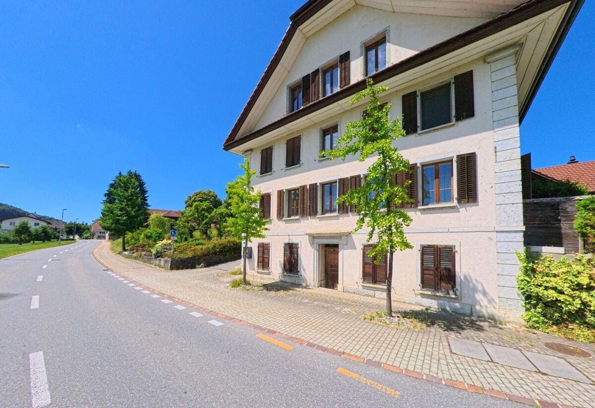 Ein mehrstöckiges Wohnhaus mit Fensterlaeden steht an einer sanft geschwungenen Strasse in Roggliswil (PLZ 6265, Kanton Luzern), umgeben von gruener Bepflanzung und Sommerlicht.