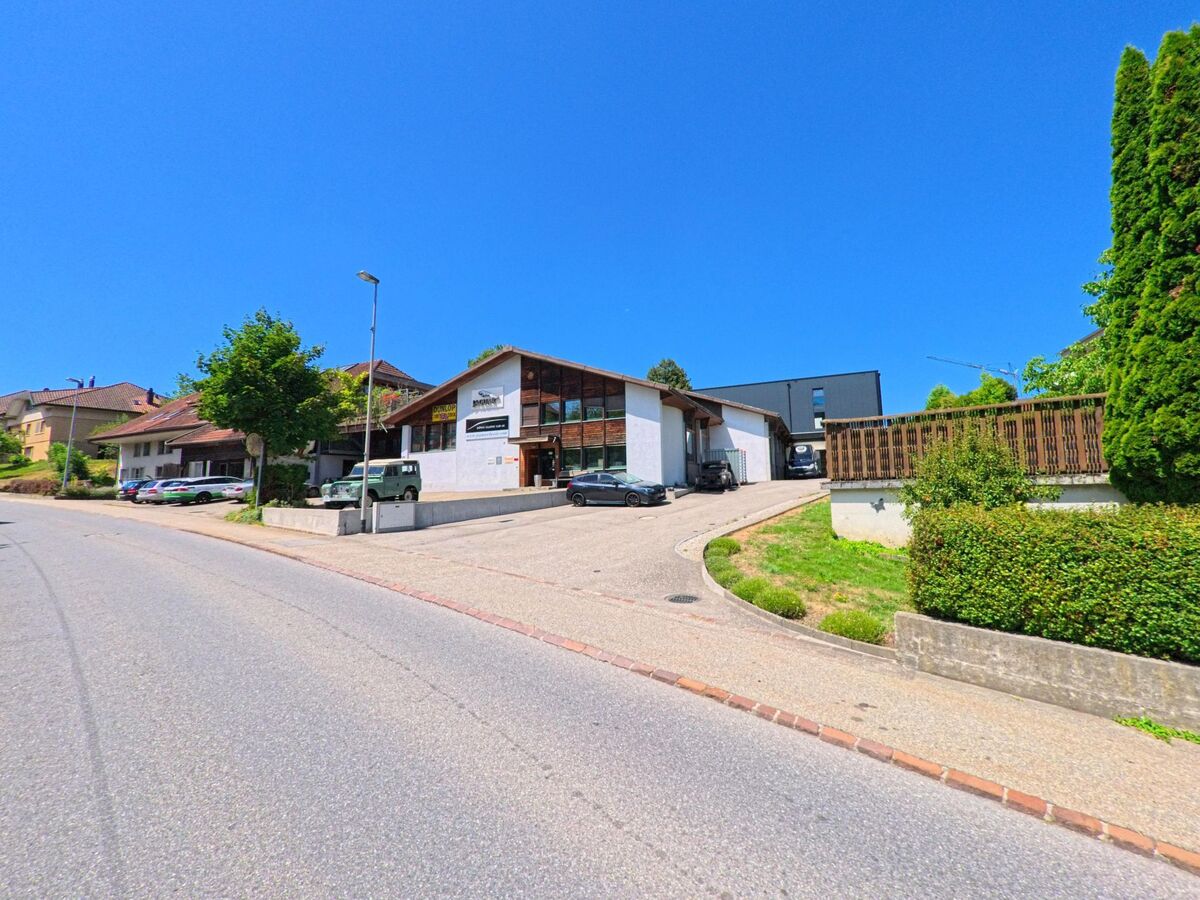 Entlang einer leicht ansteigenden Strasse in Roggliswil (PLZ 6265, Kanton Luzern) liegt ein Gewerbegebäude mit grossen Fenstern und Parkplaetzen, daneben befinden sich Wohnhaeuser und Baeume.